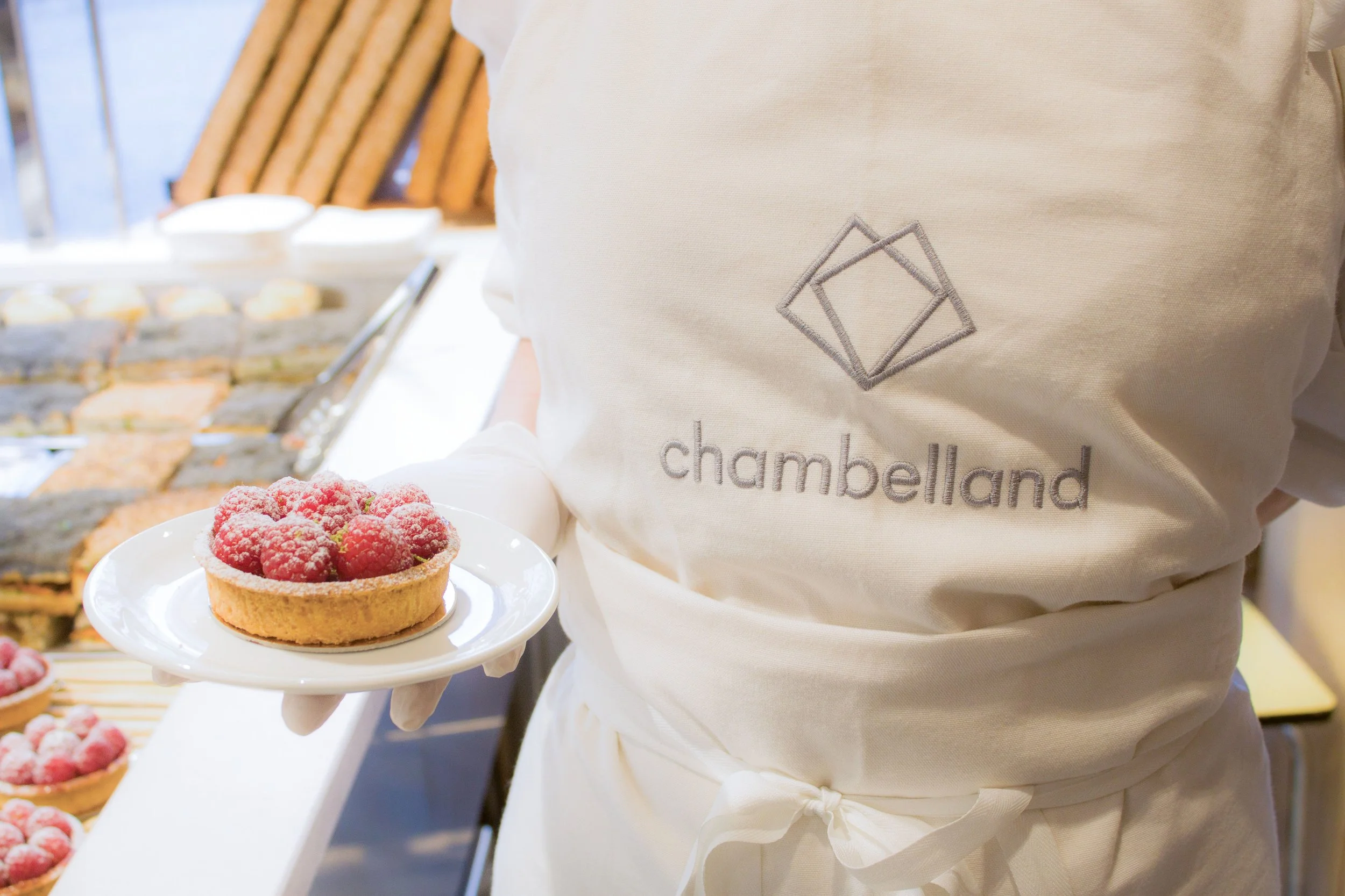 Une personne portant un tablier blanc avec le logo 'chambelland' tient une assiette avec une tarte aux framboises. En arrière-plan, il y a d'autres tartes et pâtisseries sur un comptoir.
