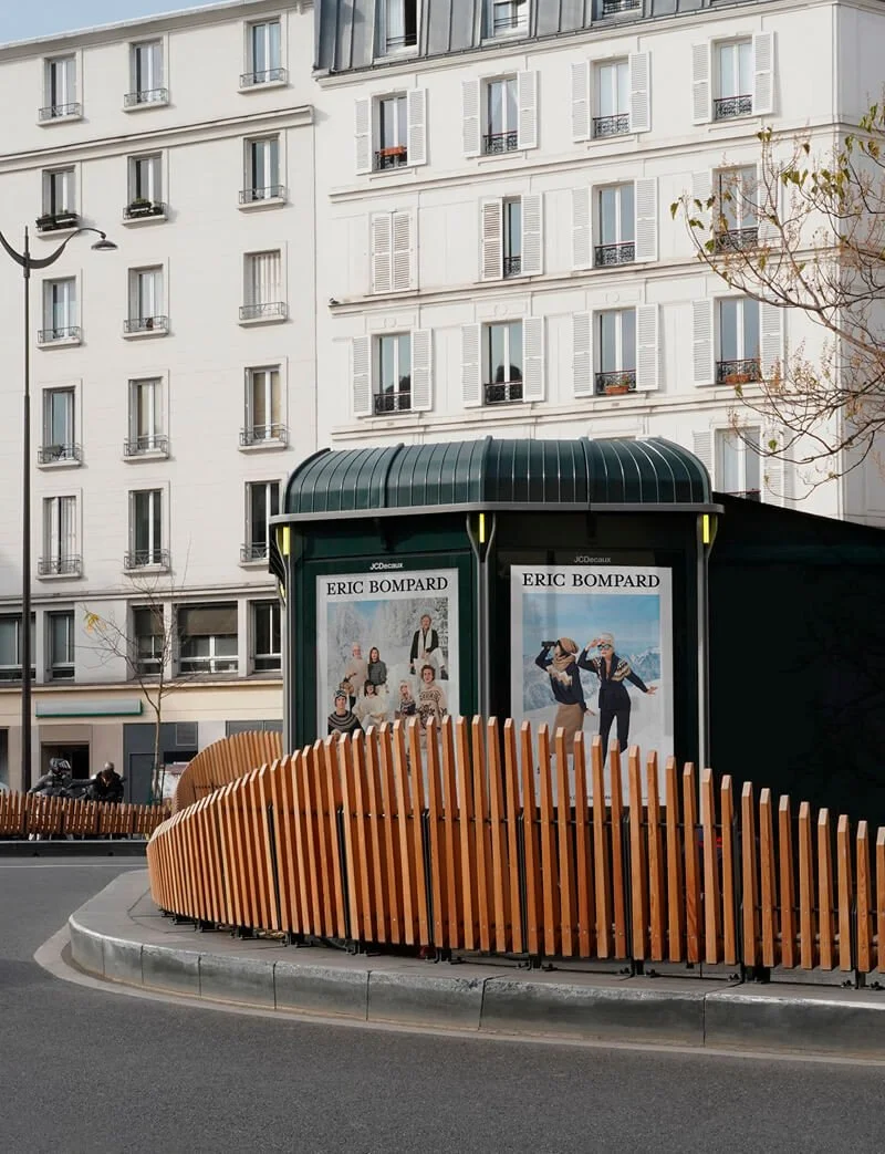 Un arrêt de bus avec une structure en bois courbe, plusieurs affiches publicitaires pour la marque Éric Bompard, et un bâtiment résidentiel en arrière-plan.