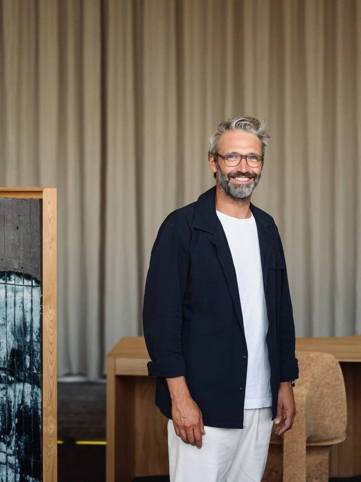 Un homme souriant portant des lunettes, une chemise noire et un t-shirt blanc, debout dans une salle avec des rideaux beige en arrière-plan.