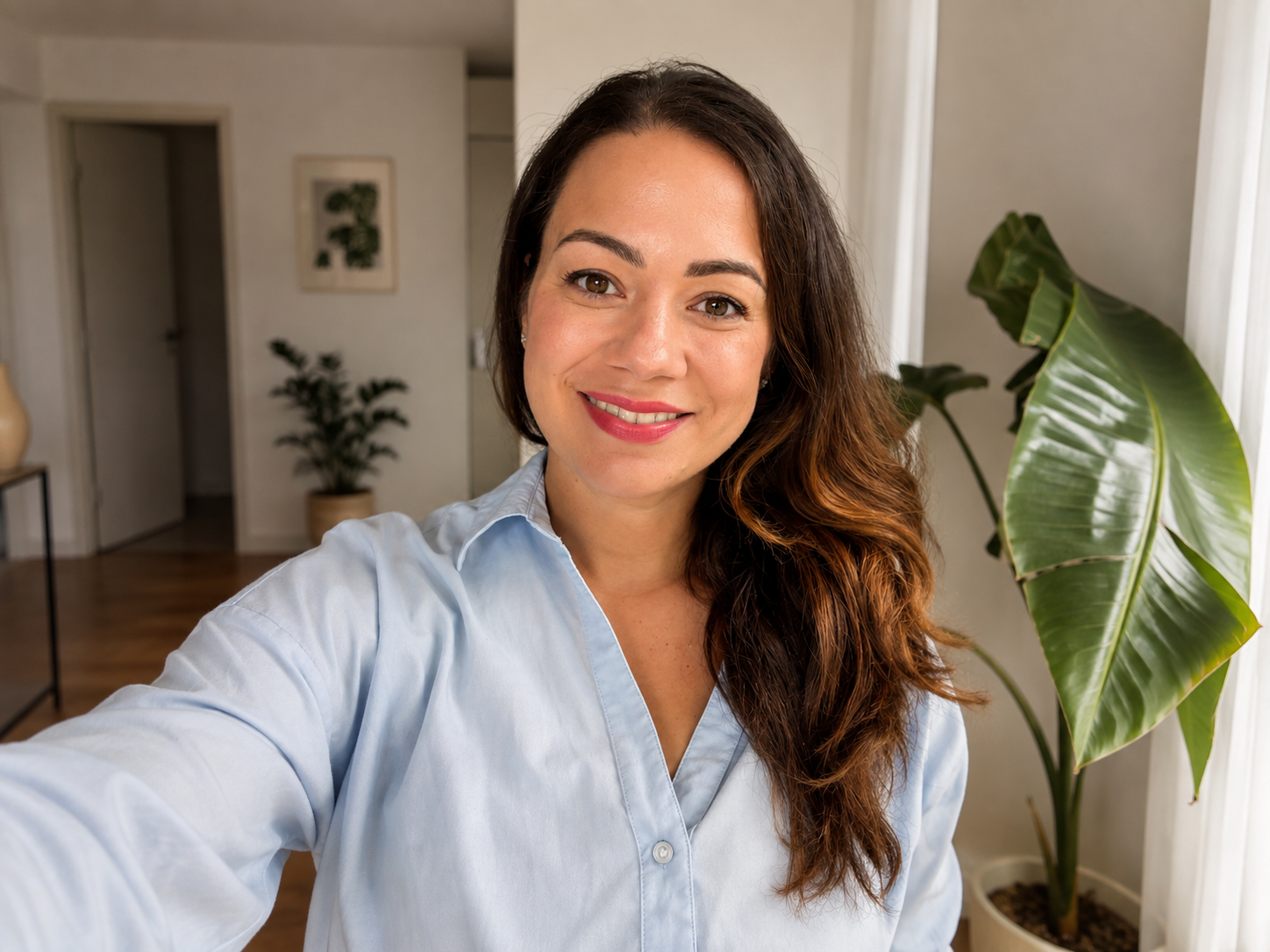 A woman with long brown hair smiling and taking a selfie indoors, with green houseplants and light-colored walls in the background.