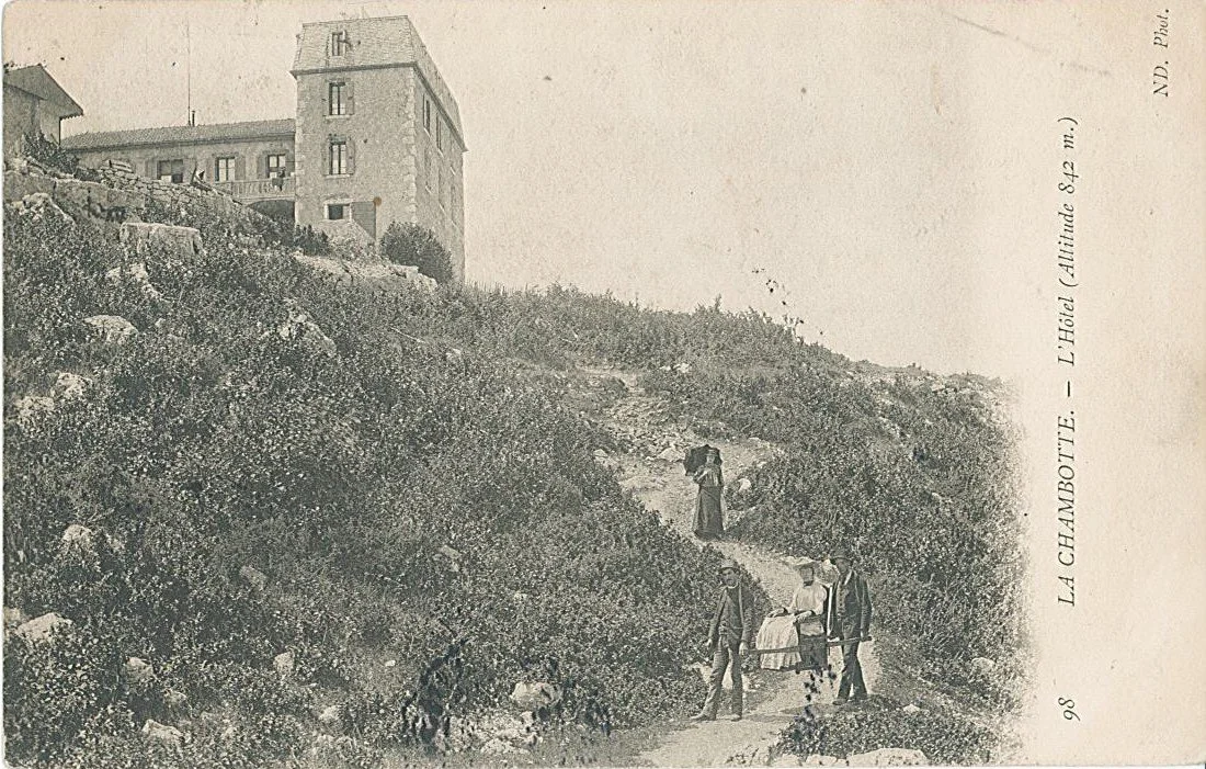 Photographie ancienne montrant un chemin montant une colline avec des personnes marchant, un chien, et un grand bâtiment au sommet de la colline, identifié comme un hôtel à La Chambotte, à une altitude de 842 mètres.