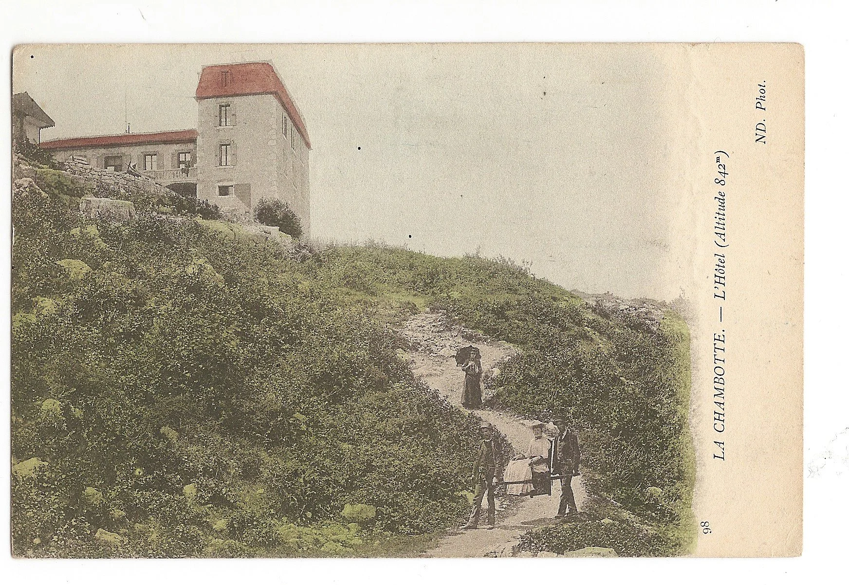 Photographie ancienne montrant un chemin entre des buissons menant à un hôtel bâti sur une colline, avec des personnes marchant et un chariot dans le chemin.