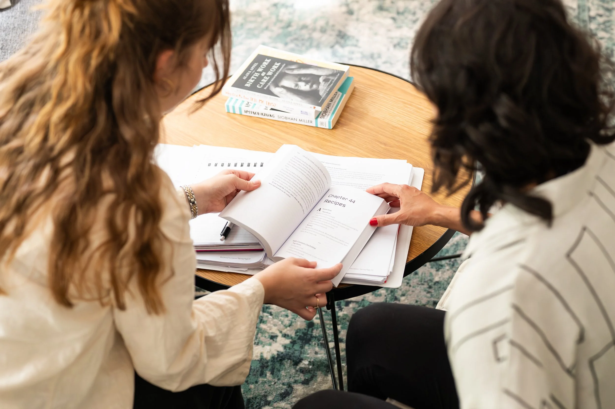 Two women sitting at a round wooden table, looking at open documents, with stacks of books nearby.