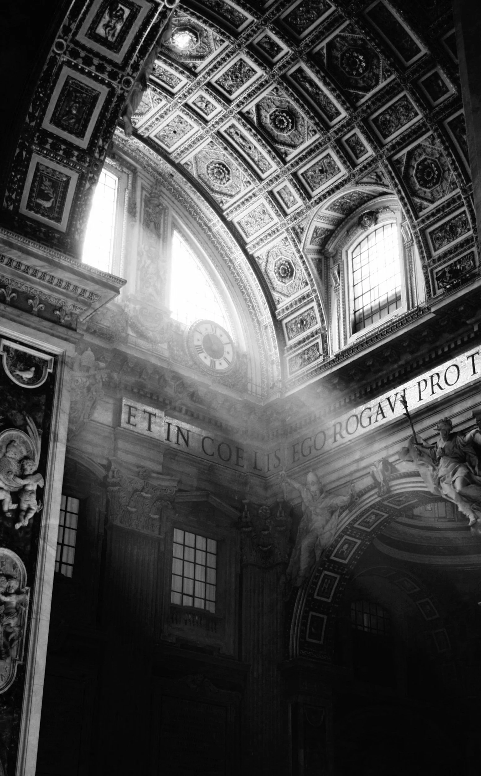 Interior view of an ornate cathedral ceiling in black and white, featuring detailed decorative patterns, an inscription in Latin, large arched windows, and sculptures of angels.