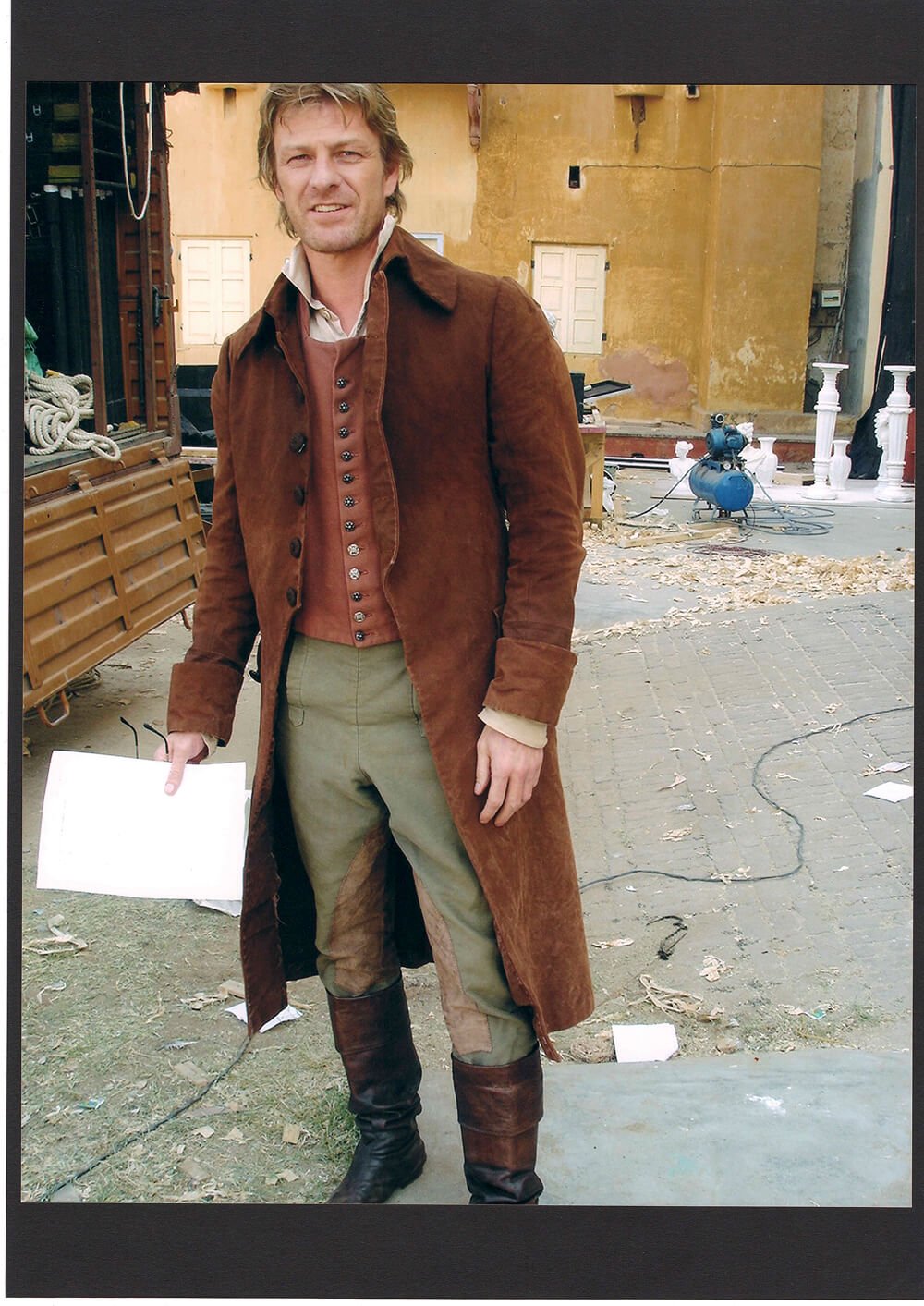 Sean Penn dressed in historical costume standing on a film set with old buildings and construction equipment in the background.
