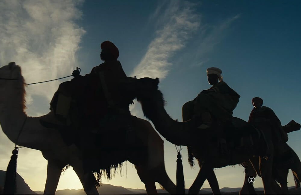 Silhouettes of two people riding camels at sunset or sunrise with a mountainous landscape in the background.