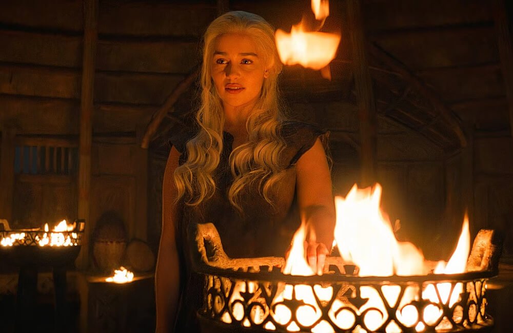 A woman with long wavy blonde hair standing in a dimly lit room near a fire. She appears to be holding or leaning on a circular fire pit, with visible flames. The room has a rustic interior with wooden walls.