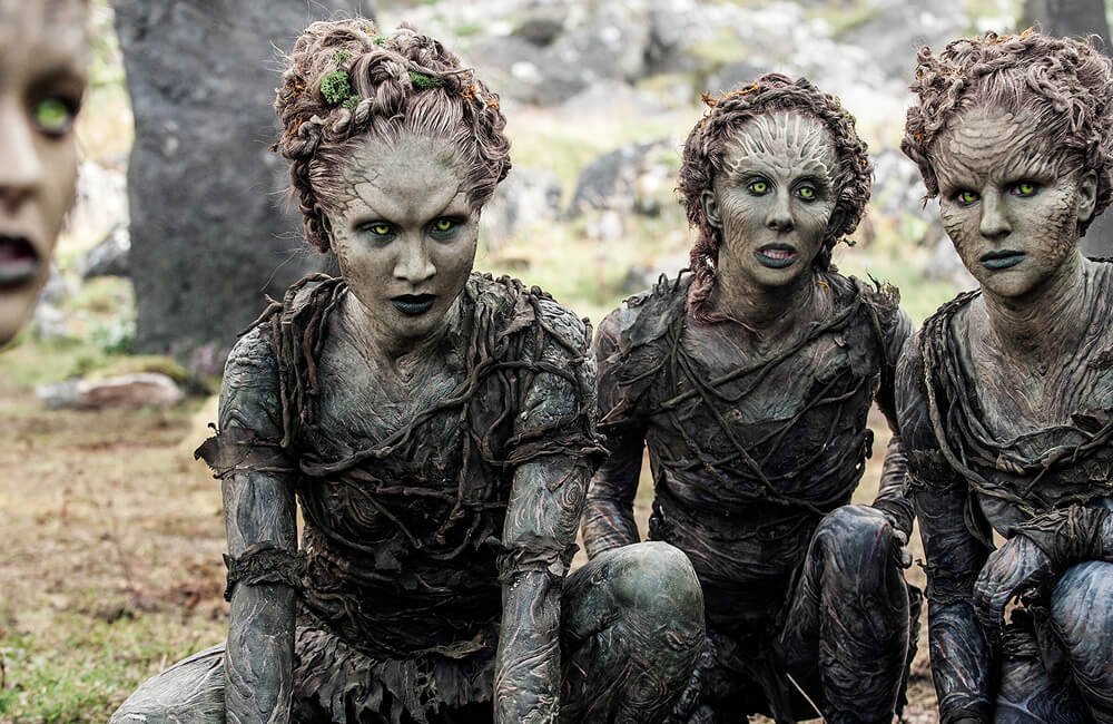 Three Children of the Forest from Game of Thrones resembling humanoid creatures with green eyes, painted faces, and textured clothing, sitting outdoors on grass with rocks and trees in the background.
