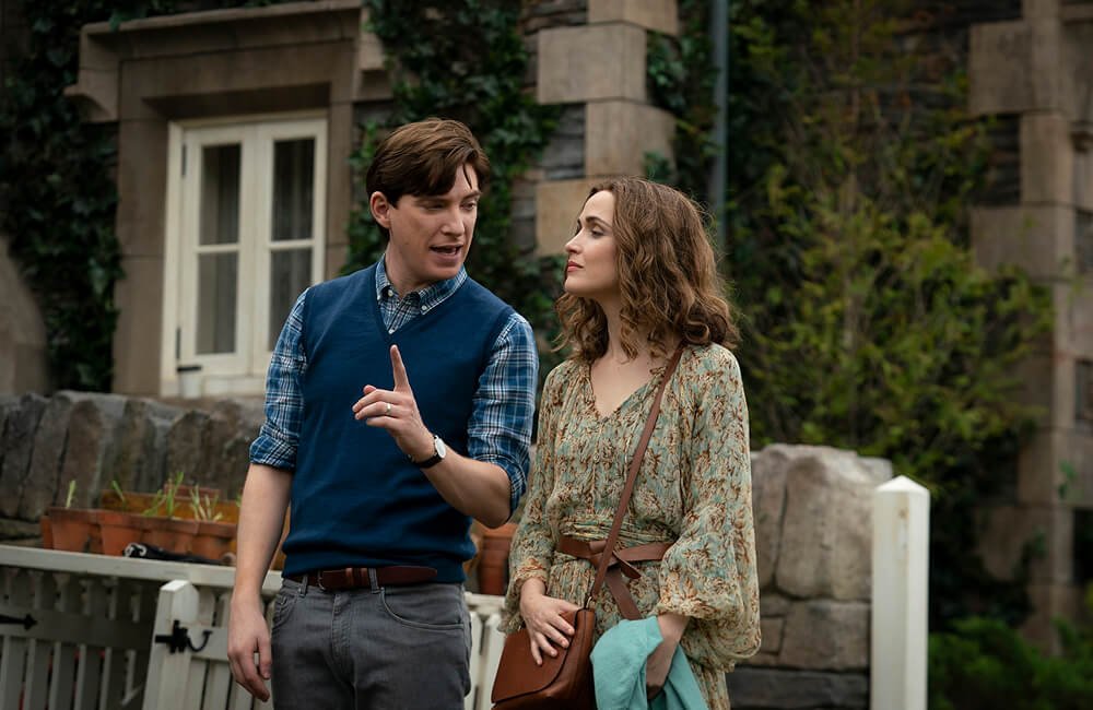 Domhnall Gleeson and Rose Byrne are having a conversation outdoors in front of a stone building with ivy in Peter Rabbit 2. The man is gesturing with his finger, and the woman is listening attentively.