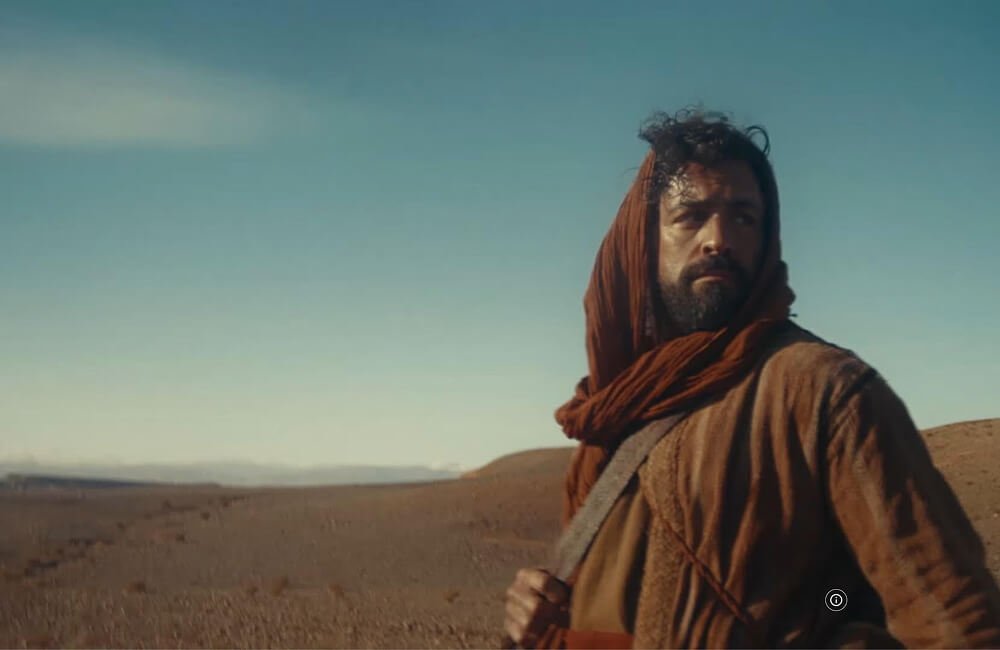 A man with a beard wearing a brown scarf, jacket, and carrying a bag, standing in a desert landscape with a clear blue sky in Jesus Crown of Thorns.