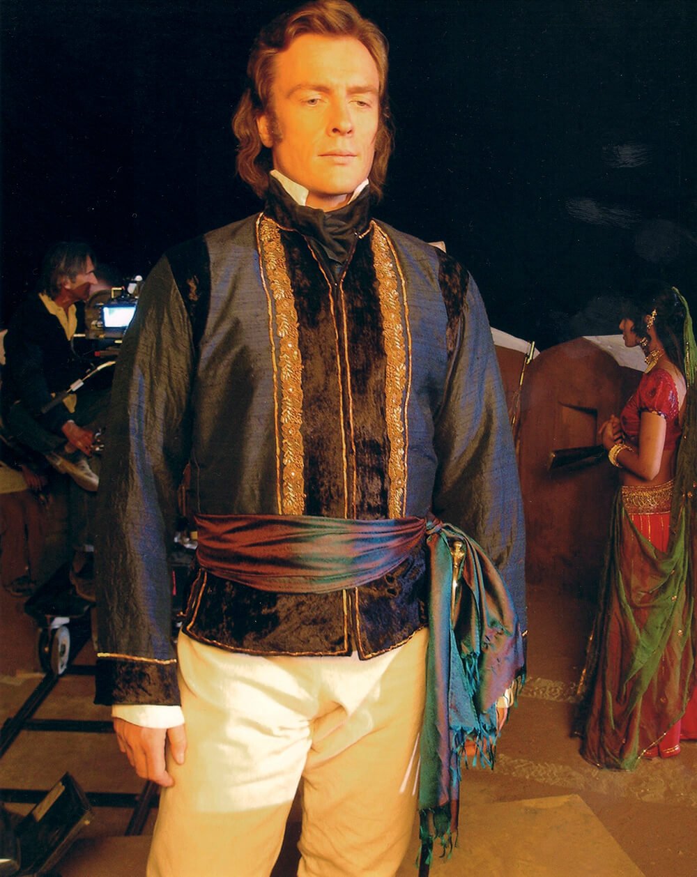Toby Stephens dressed as a historical figure in a dark, ornate jacket with gold embroidery, cream-colored pants, and a colorful sash, standing on a film set with a woman in traditional Indian attire and a crew member in the background.