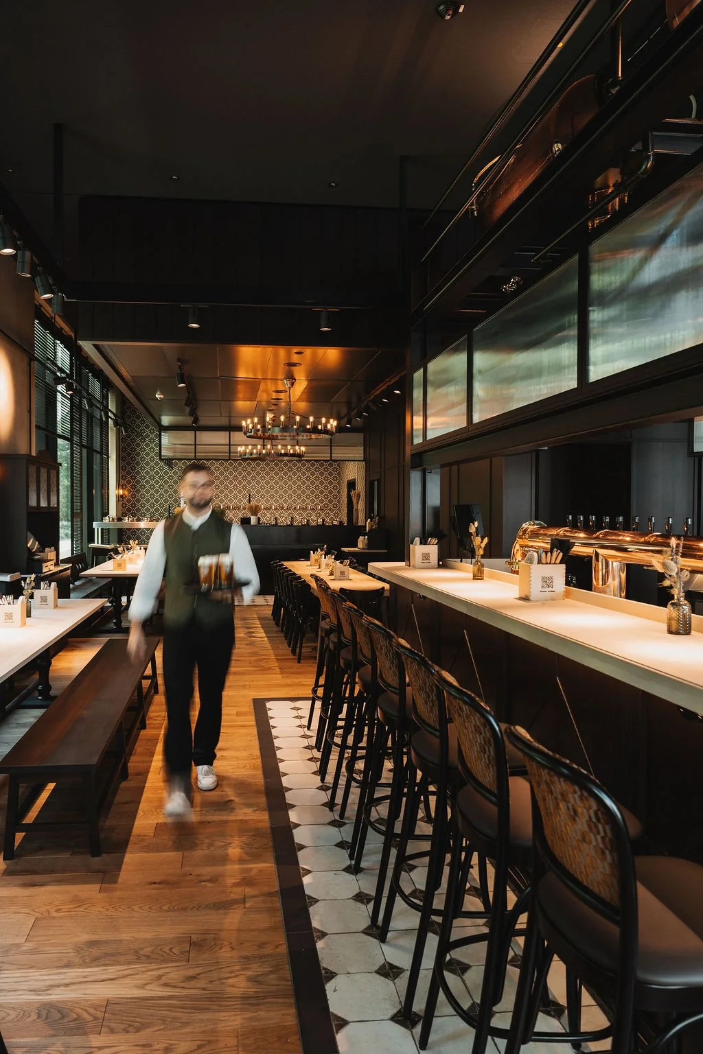 Interior of a modern restaurant with wooden floors, bar seating, and a server carrying drinks, illuminated by chandeliers.