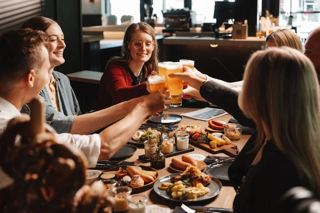 Group of friends enjoying a meal and toasting with glasses of beer at Schwabinger Tor Bräu Wirtshaus Stammtisch table filled with various local dishes.