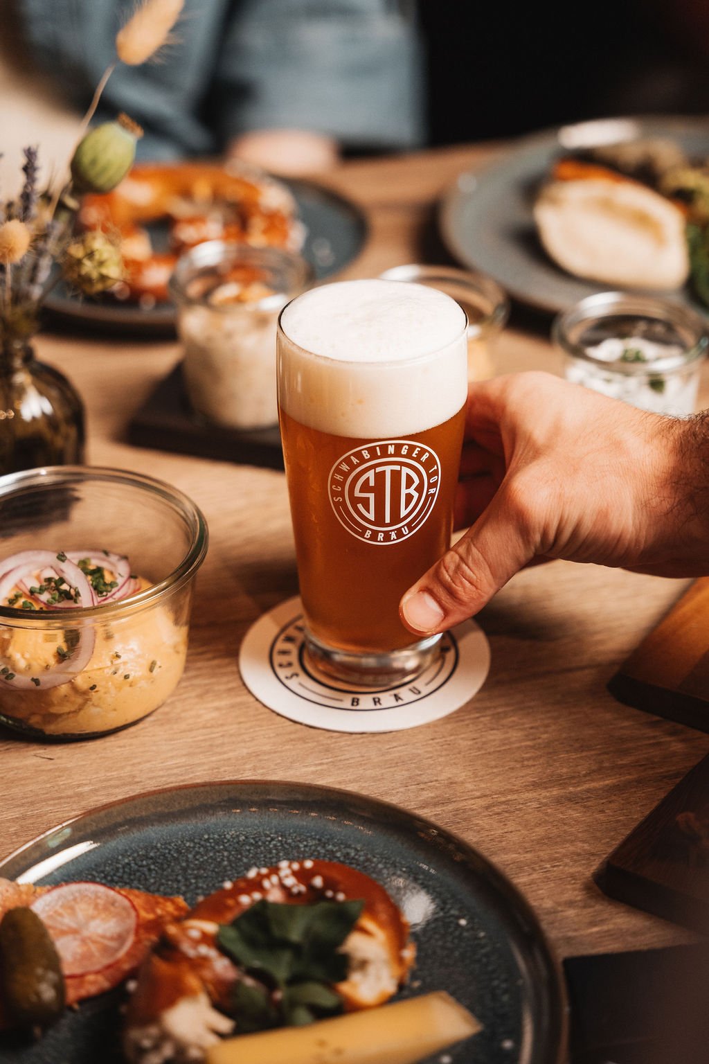 A person holding a glass of beer with a foam head at a wooden table with various dishes and small bowls of food.