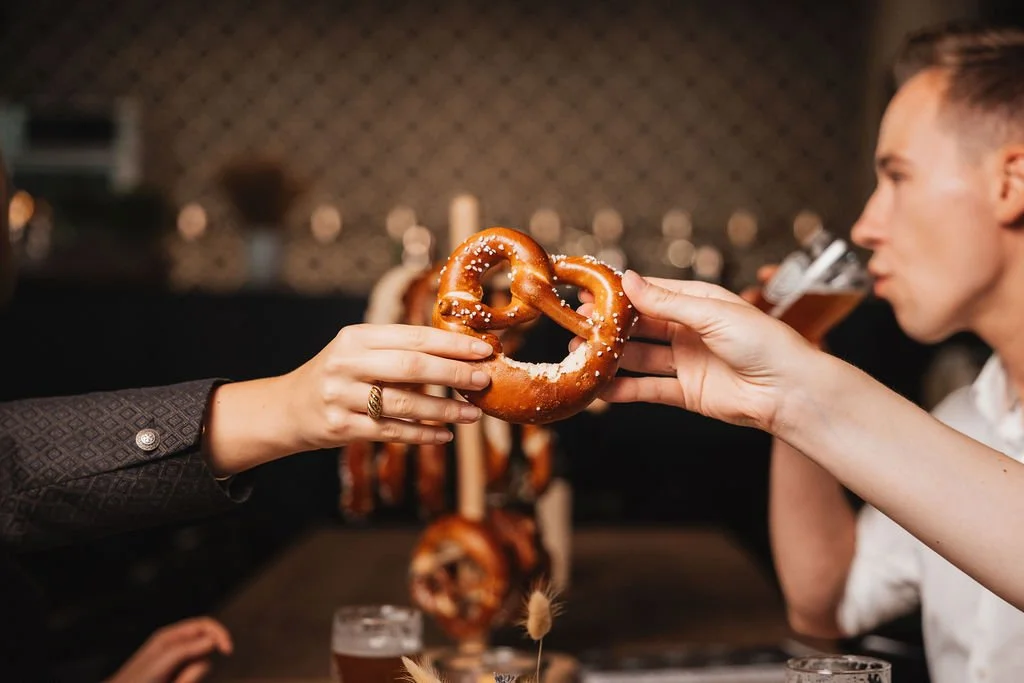 Two people exchanging a pretzel at a social gathering with drinks on the table.