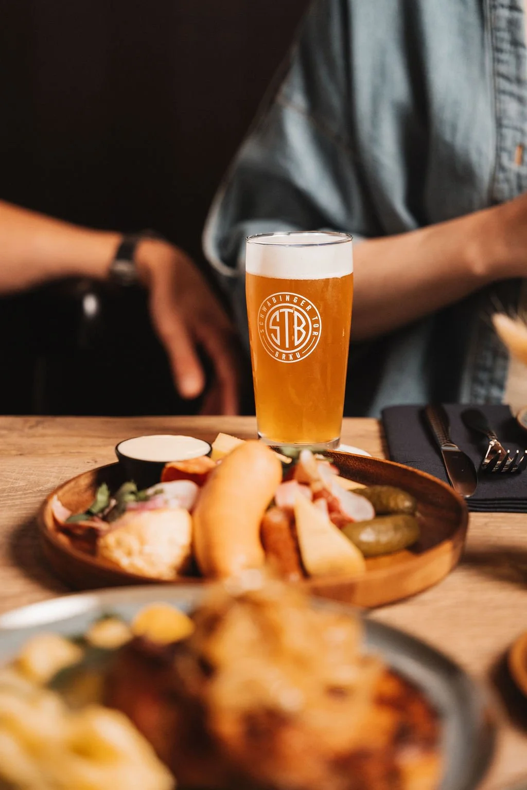 A glass of beer on a wooden table with pub food including a hot dog, pickles, and fries.