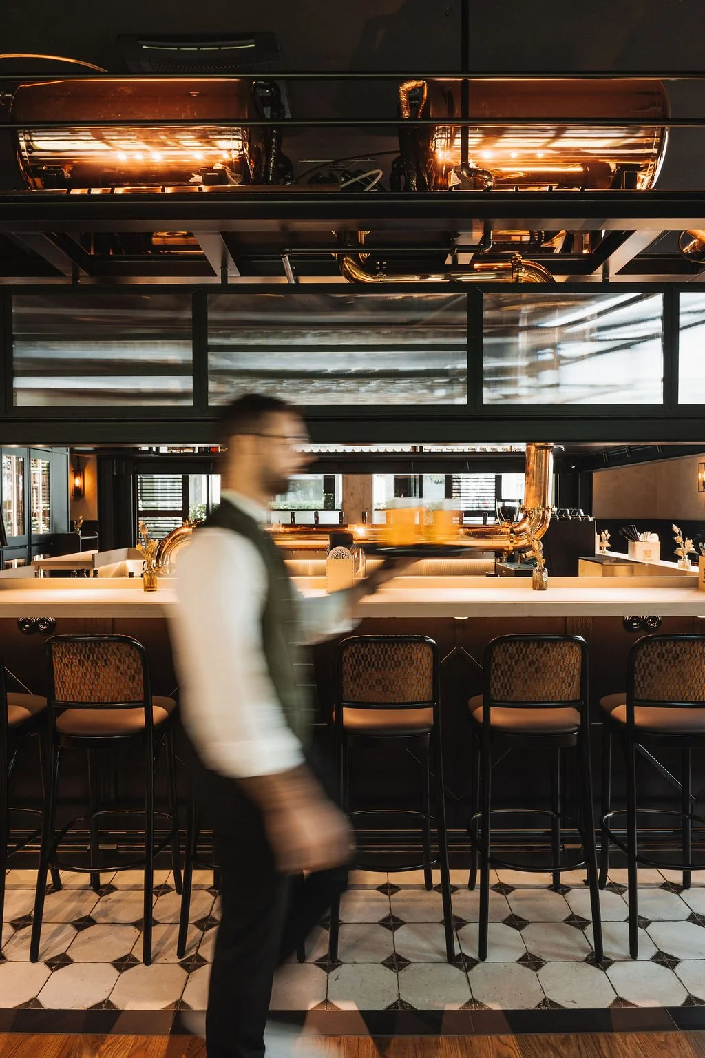 A blurred waiter in a white shirt and black vest carrying a tray with two drinks inside a modern restaurant with bar stools, a white bar counter, and industrial-style decor.