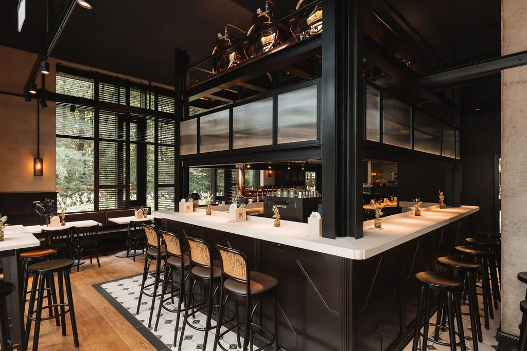Interior of a modern restaurant with black accents, white marble bar counter, bar stools, and large windows with blinds.