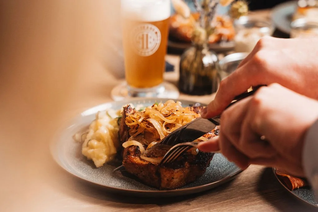 Person cutting into a serving of steak topped with fried onions, served with mashed potatoes on a gray plate. In the background, there is a glass of beer and a small vase with flowers on the table.