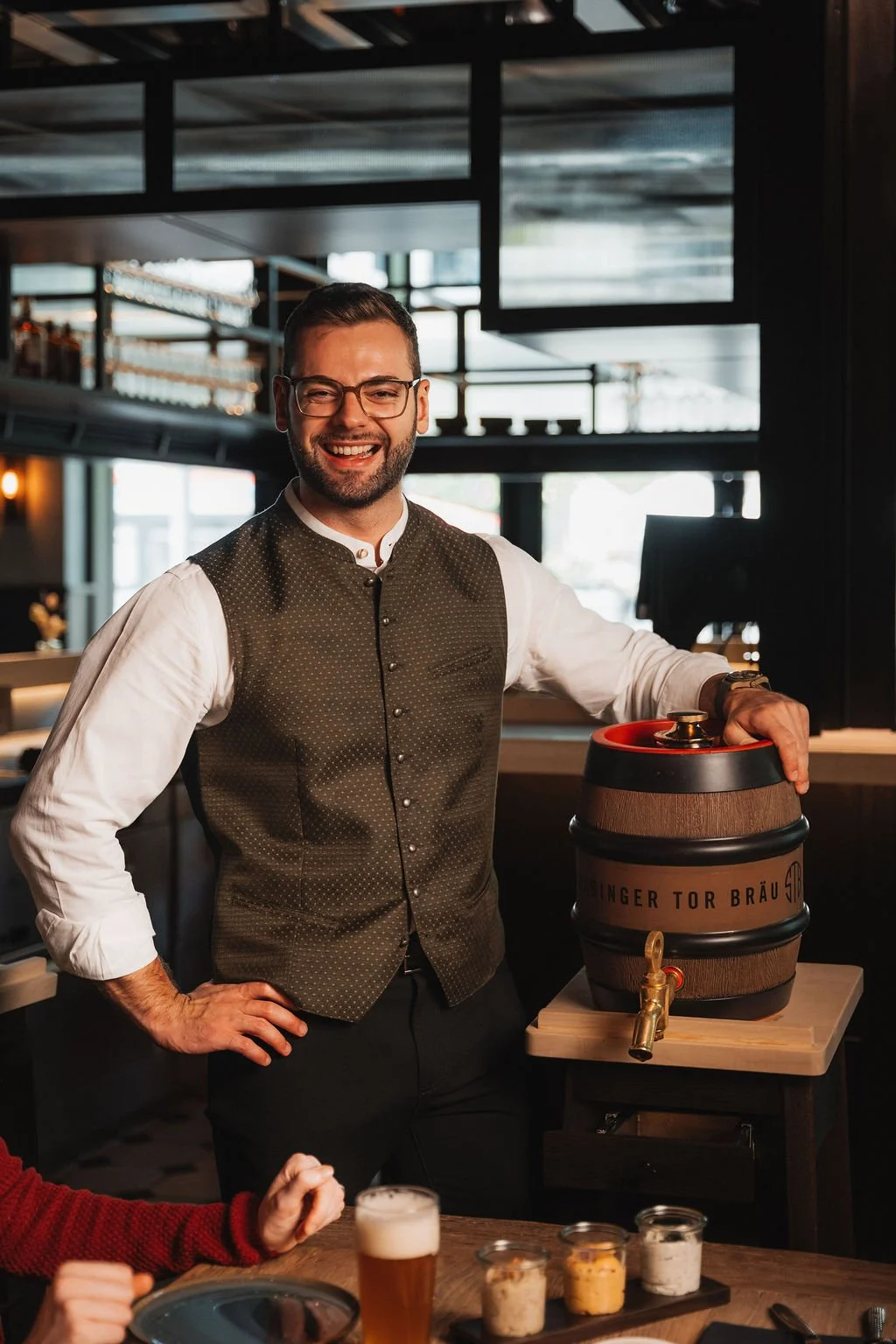 Wirt Johannes Egger in a traditional Bavarian outfit standing in a restaurant, smiling, with a beer tap and a beer barrel labeled 'Schwabinger Tor Bräu' beside him.
