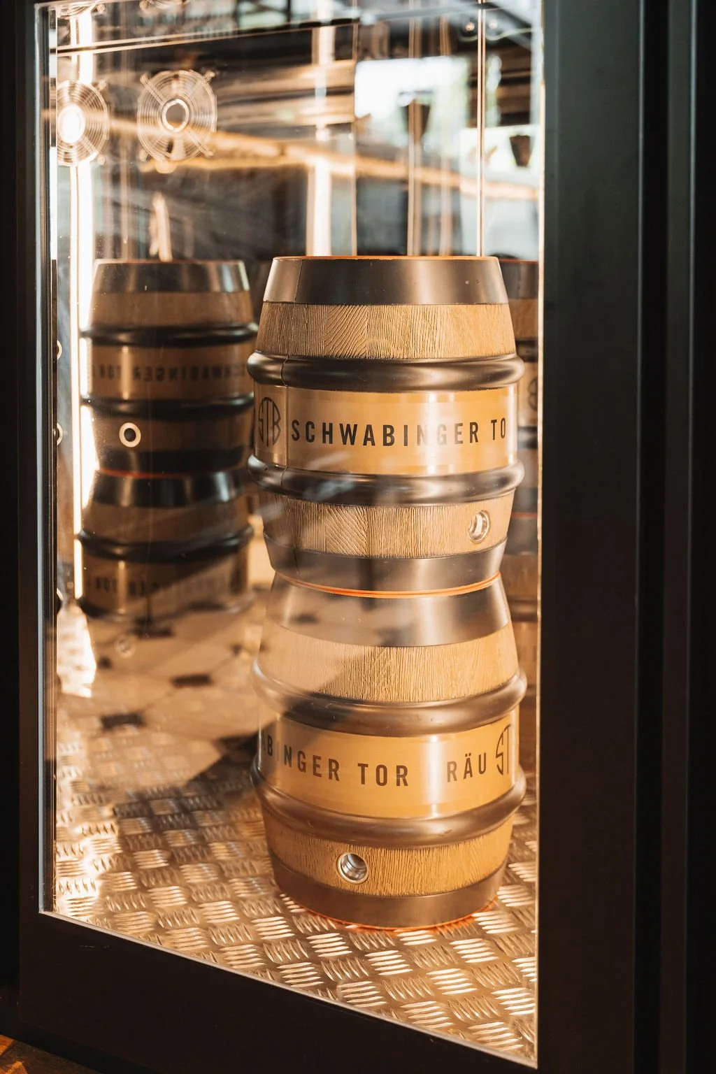 Stacked wooden and metal barrels inside a glass display case, with visible text "Schwabinger Tor" on the front barrel.