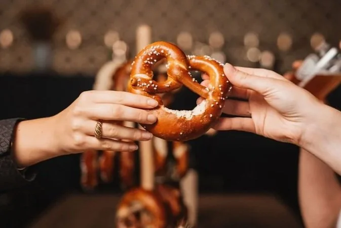 Two people are exchanging a large pretzel, with one person holding it from each side, in front of a background with additional pretzels hanging.