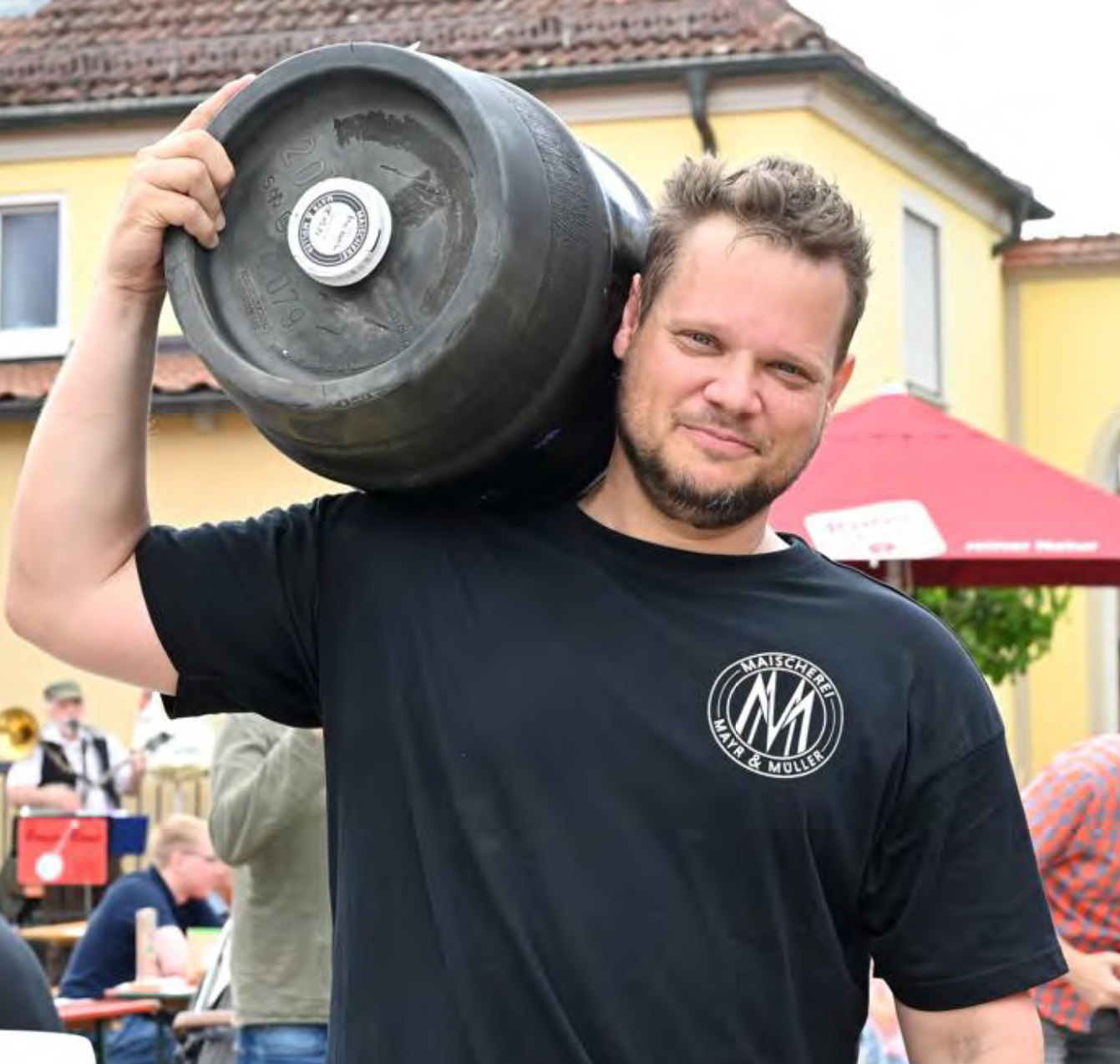 A man with a beard and short hair carries a large black weight on his shoulder outdoors, with people and buildings in the background.