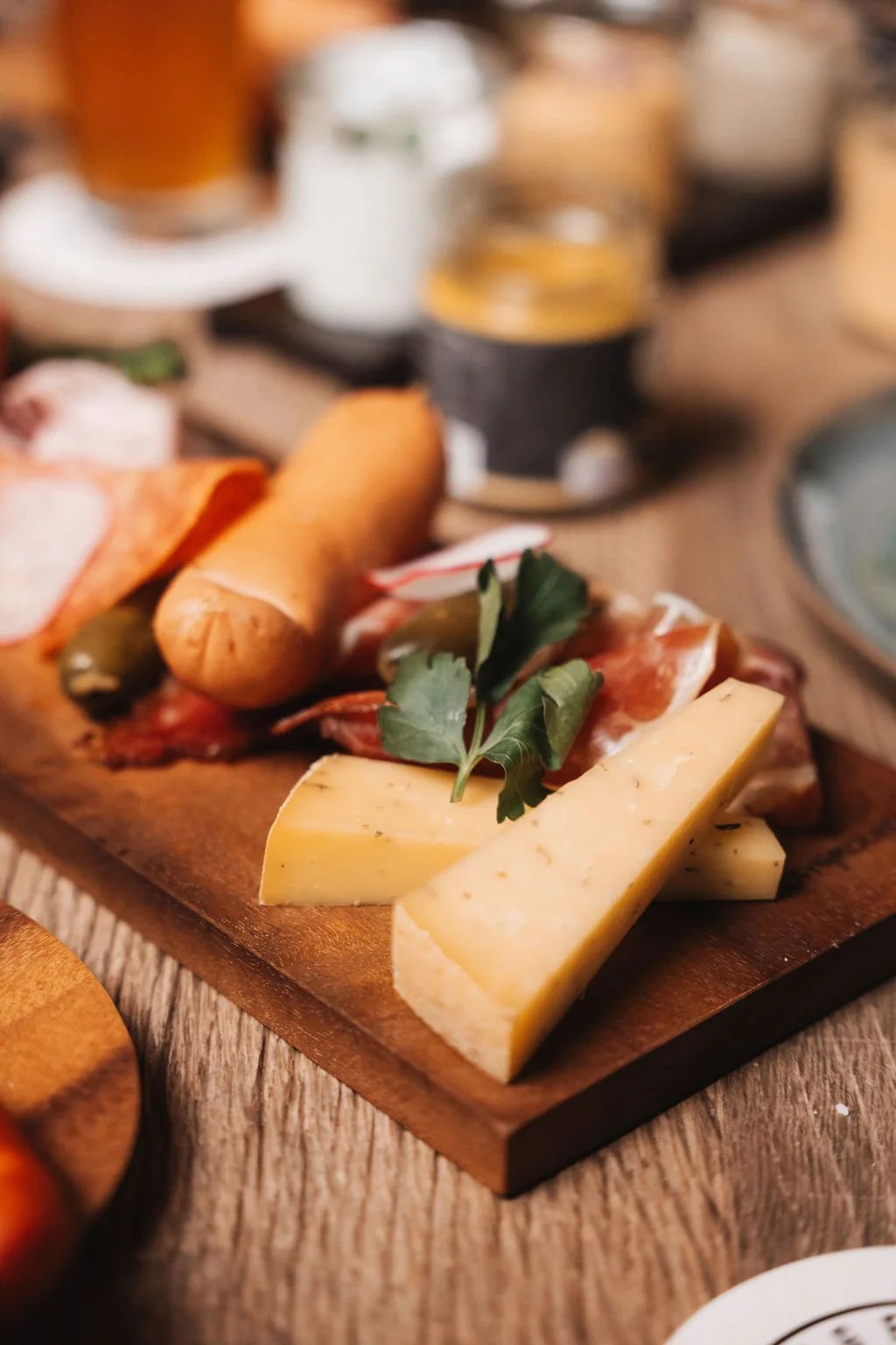 Close-up of a cheese and meat platter with herbs on a wooden board, with drinks in the background.