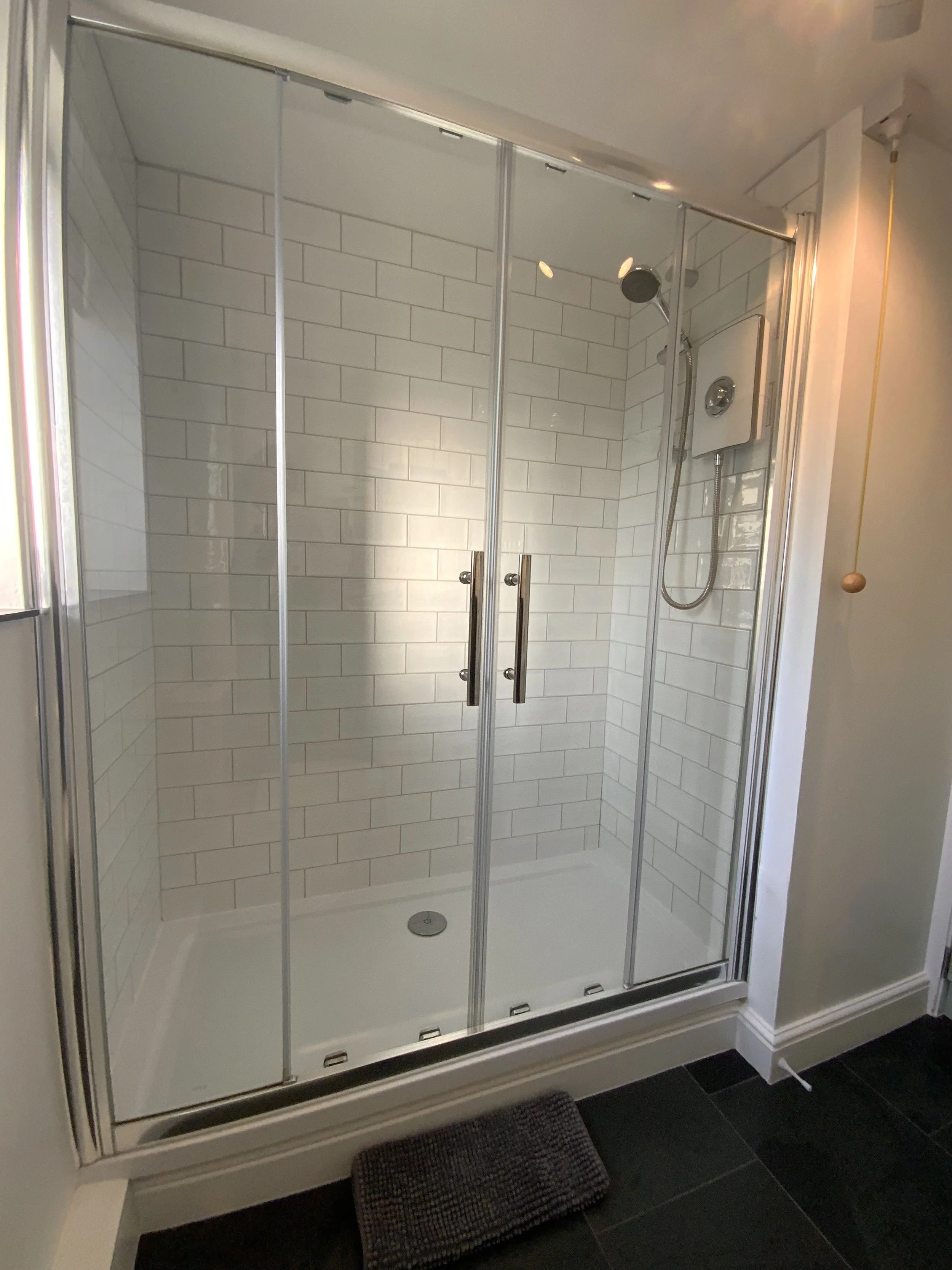 Modern shower enclosure with glass doors, white subway tiles, chrome showerhead, and gray mat on black tiled floor.