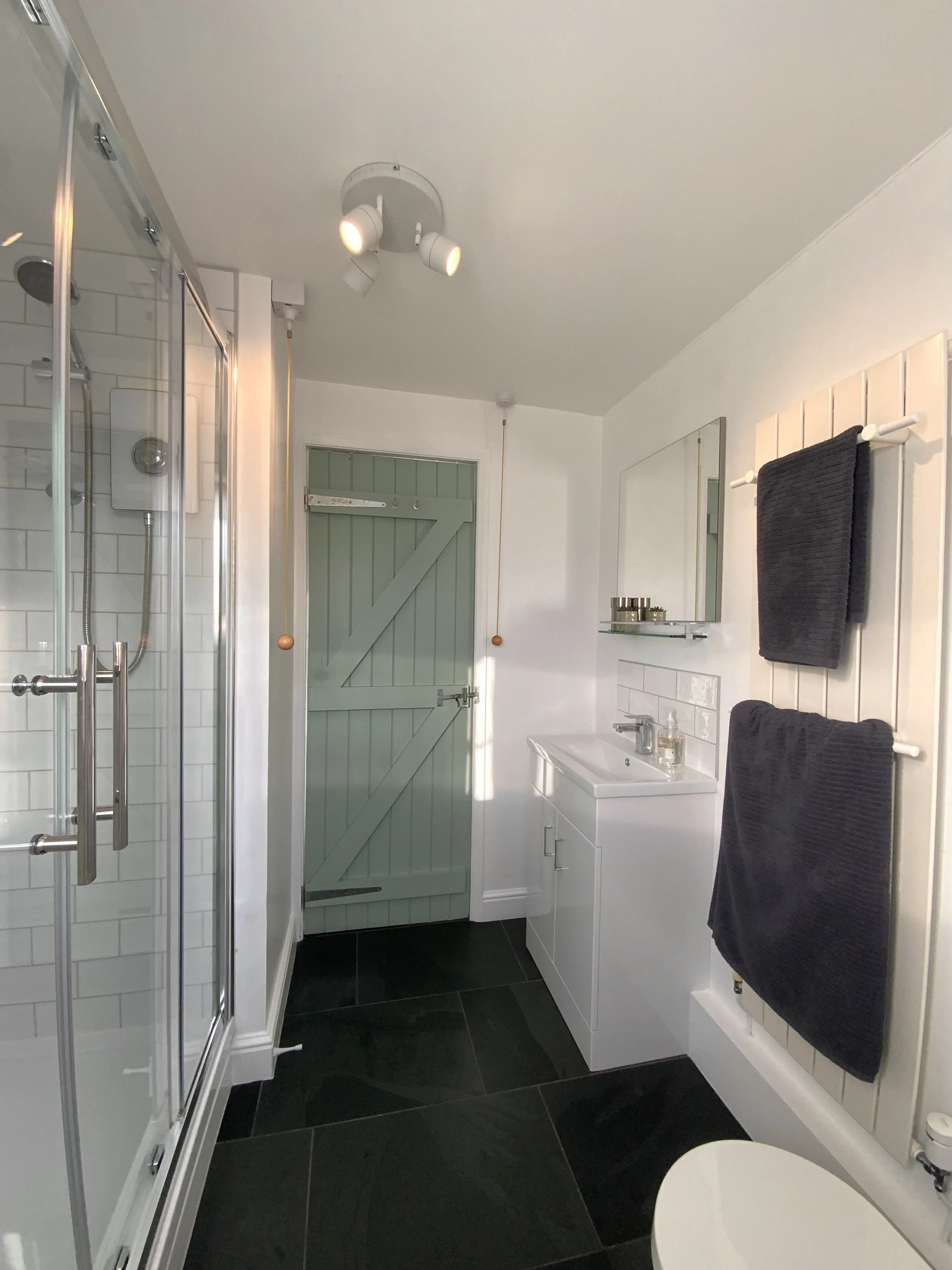 Modern bathroom with glass shower, mint green door, white vanity with sink and mirror, black tiled floor, and dark towels on a rack.