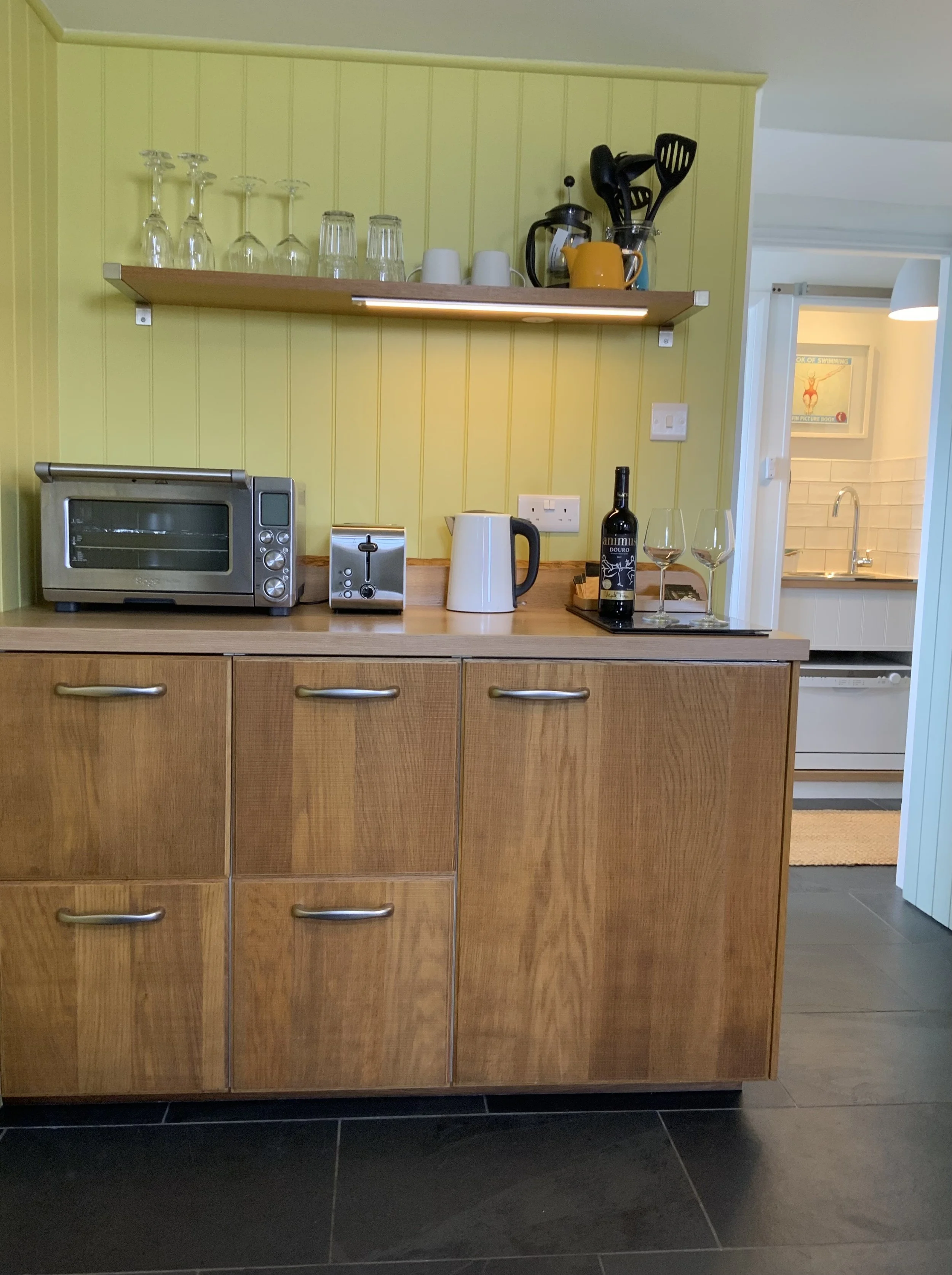 Kitchen area with a yellow wall, wooden cabinets, a shelf with glasses, a toaster oven, kettle, toaster, utensils, wine bottle, and glasses.