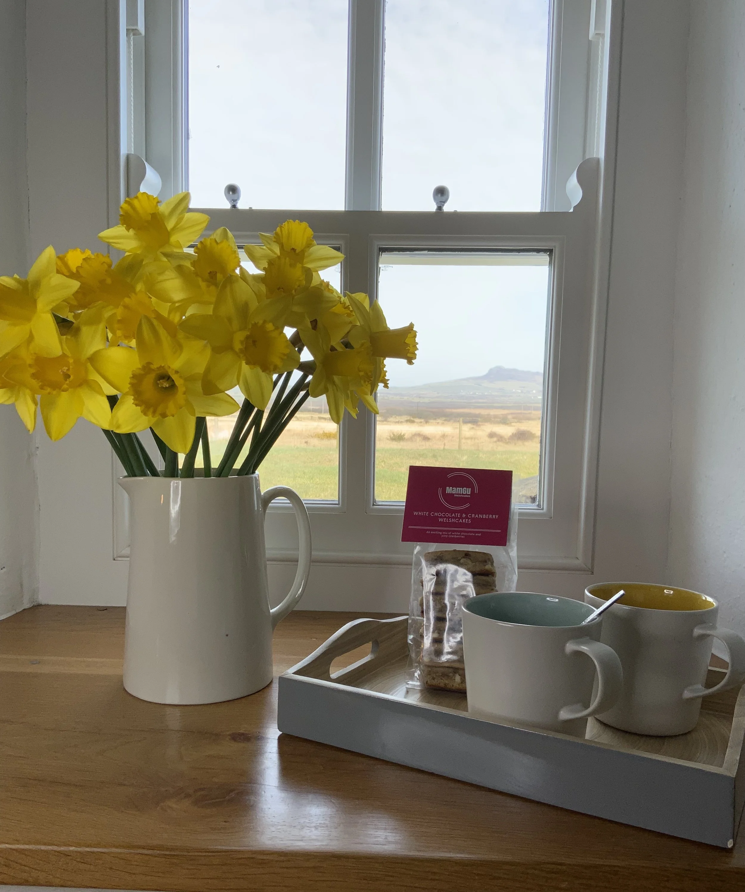 Yellow daffodils in a white pitcher on a windowsill beside a tray with two mugs, cookies in packaging, and a spoon.