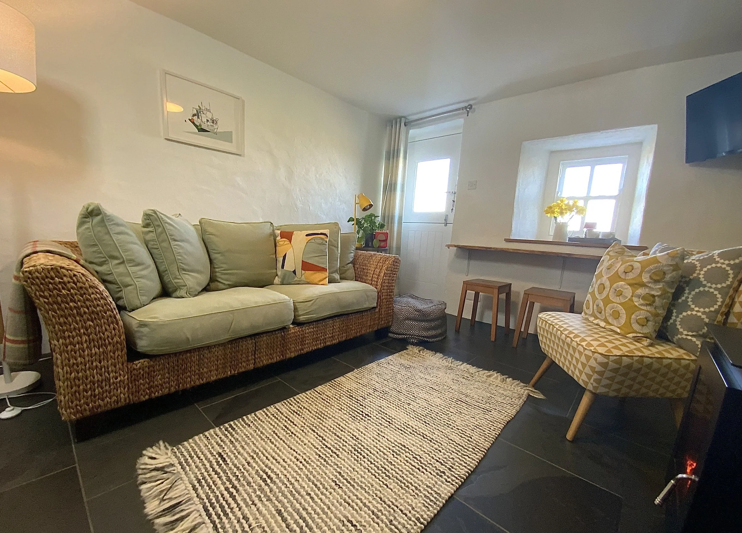 Cozy living room with a wicker sofa, patterned cushions, and a light-colored rug on dark floor tiles. There's a small cushioned chair, a window with sunlight, a wall shelf with stools, and a wall-mounted TV.