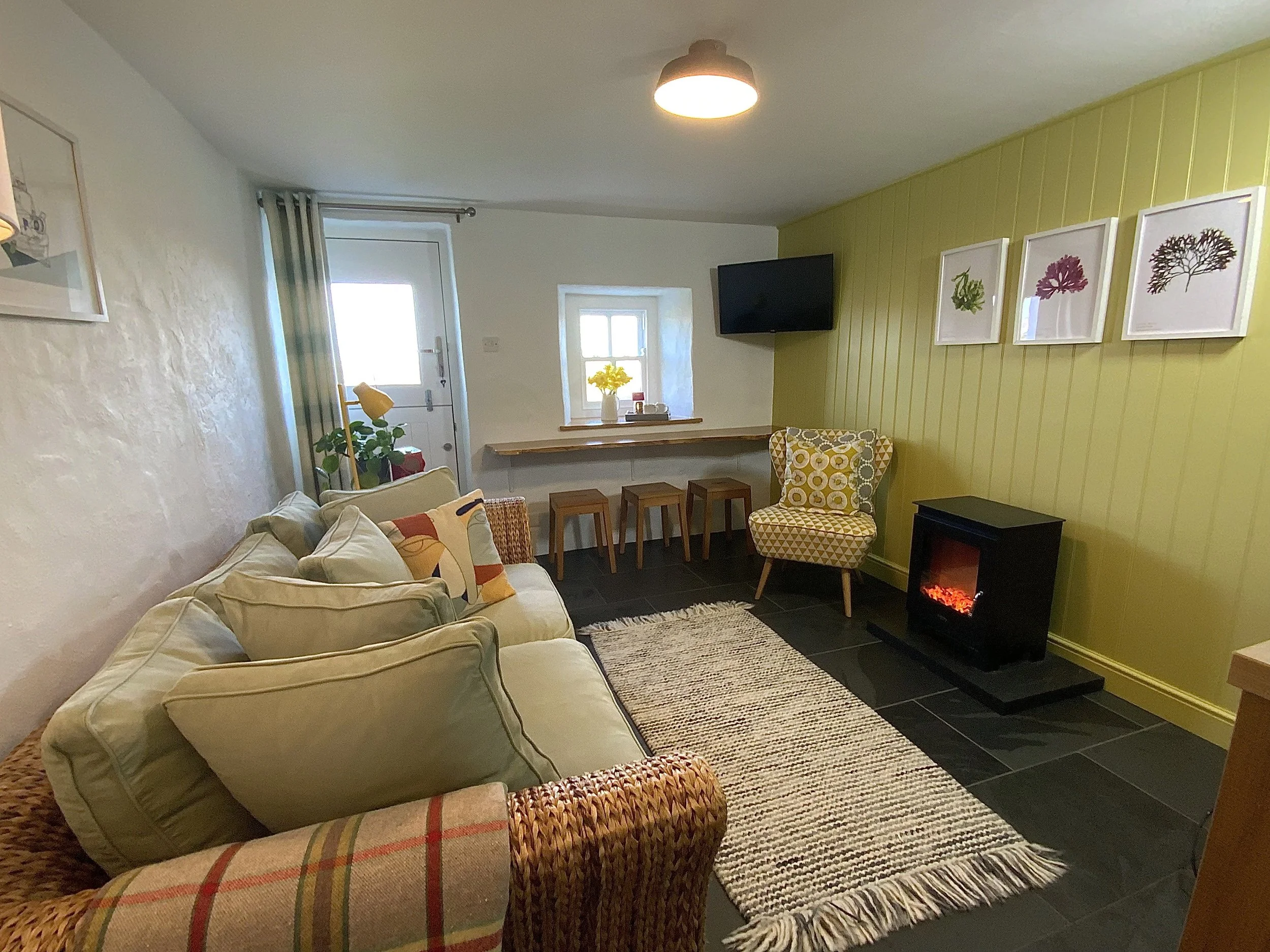 Cozy living room with a green accent wall, cream sofa, patterned armchair, electric fireplace, and small dining area. Art prints, a television, and houseplants are also present.