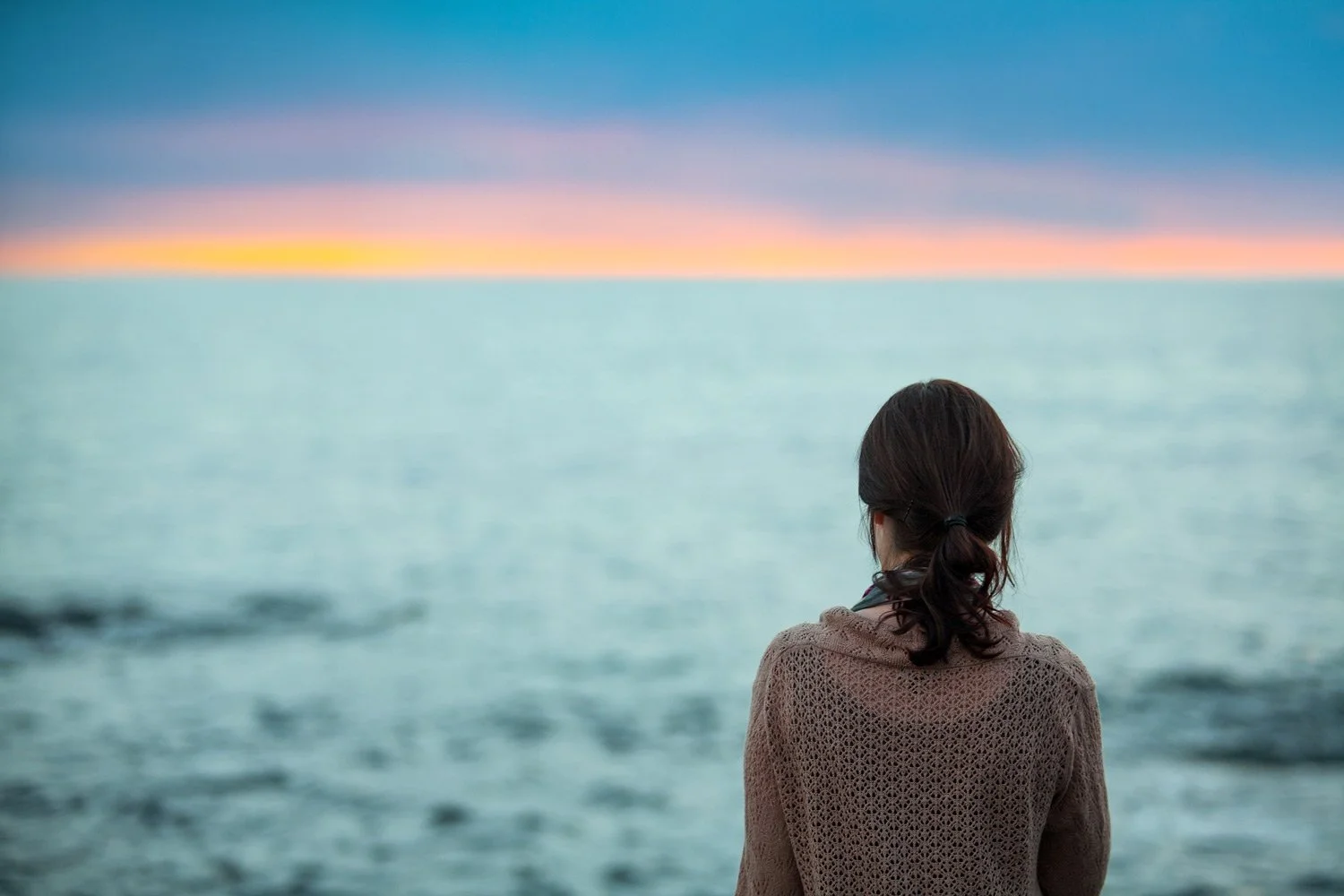 A woman with dark hair in a ponytail, wearing a beige sweater, looking out at the ocean during a sunset or sunrise with colorful sky and calm waters.