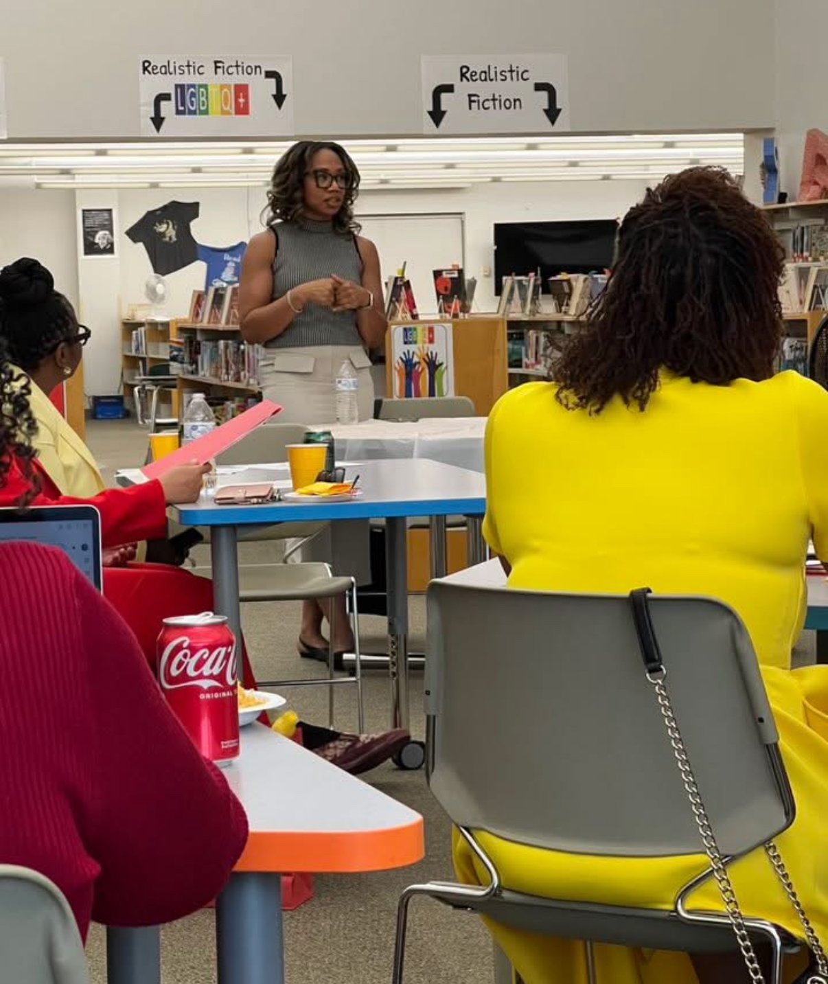 A woman standing and speaking in front of a group in a library, with a sign overhead indicating sections for 'Realistic Fiction' and 'LGBTQ+' books.