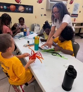 A woman with glasses teaching a craft activity to four children sitting at a table in a classroom decorated with sports-themed wall art, with supplies like scissors, paper, and water bottles on the table.