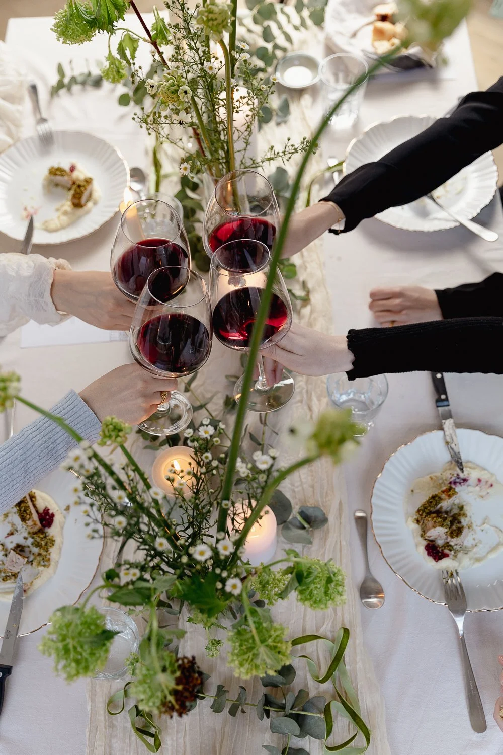 People raising glasses of red wine at a decorated dinner table with floral centerpieces and candles.