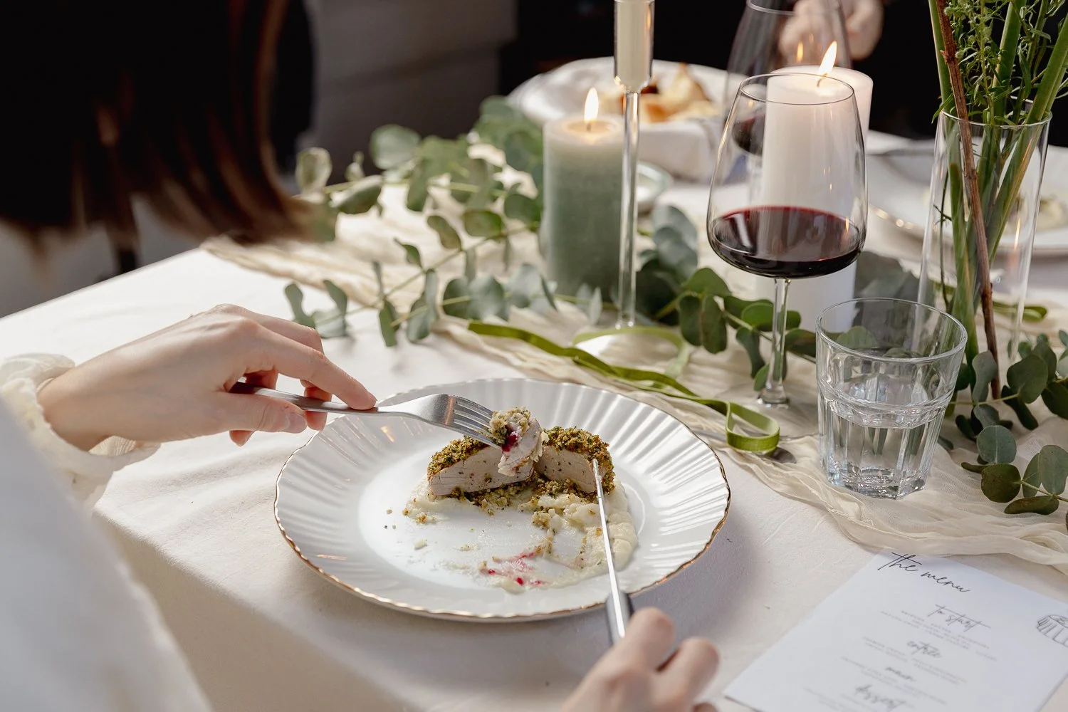 A person holding a fork and knife cutting a slice of dessert on a white plate. The table includes a glass of red wine, a glass of water, lit candles, and a menu with greenery decorations.