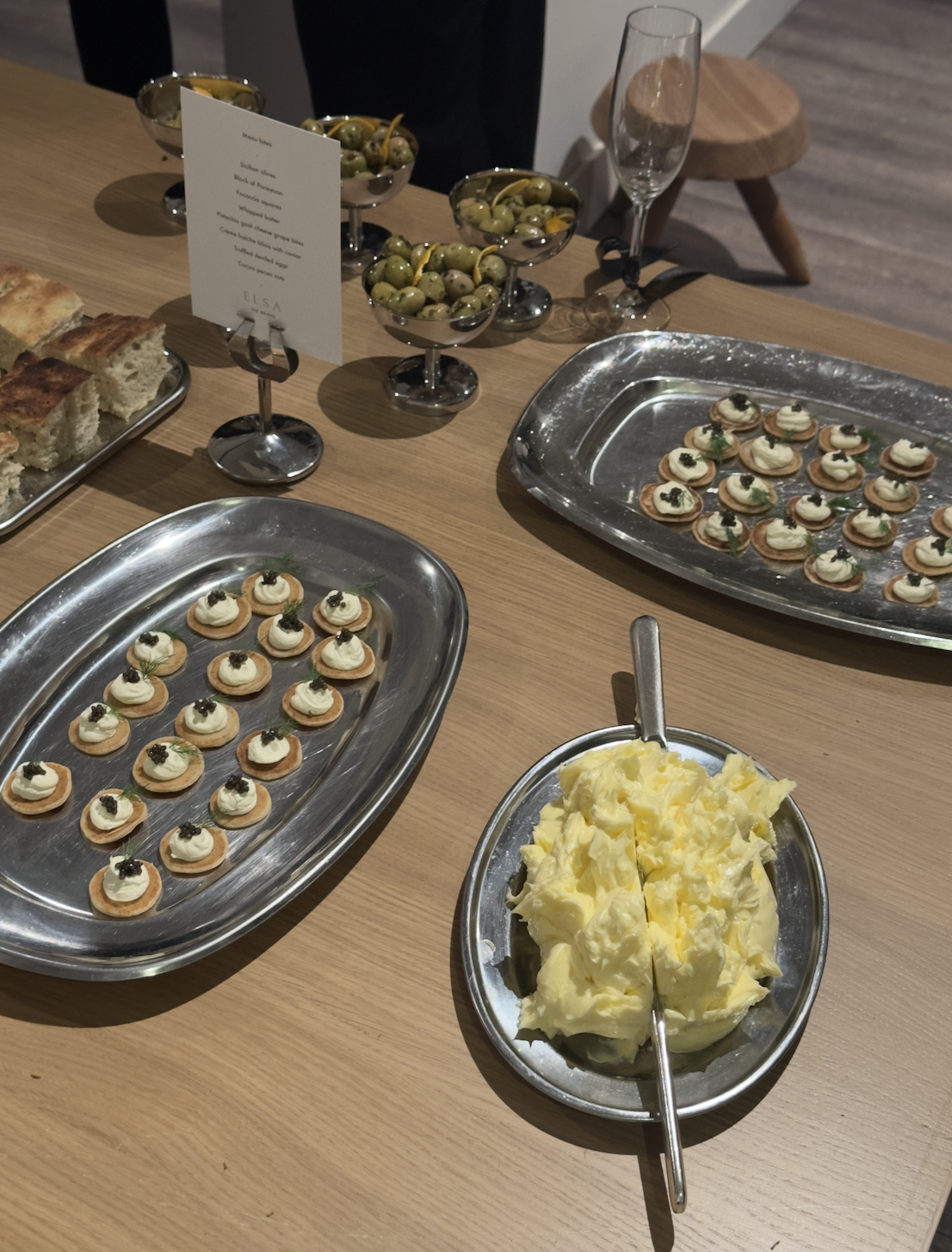Small round crackers topped with cream and black caviar on silver trays, a bowl of mashed potatoes, bowls of green olives, a glass of empty champagne, and a menu card on a wooden table.