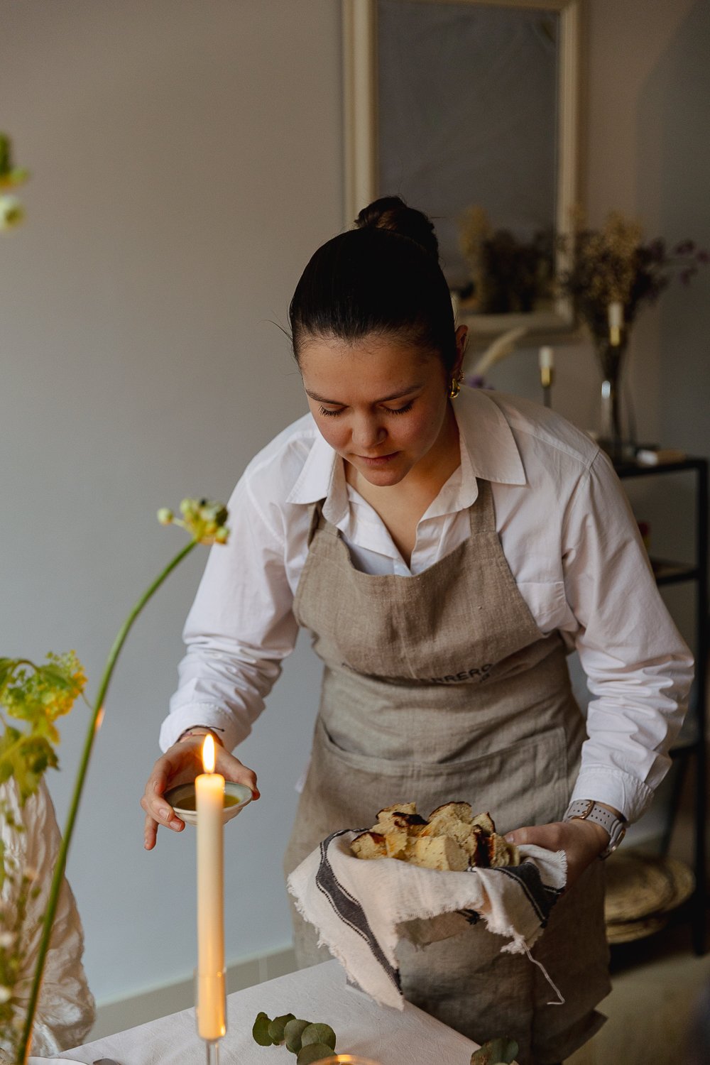 A woman wearing a white shirt and beige apron is placing slices of cake onto a white tablecloth, with lit candle nearby and a vase with flowers in the background.
