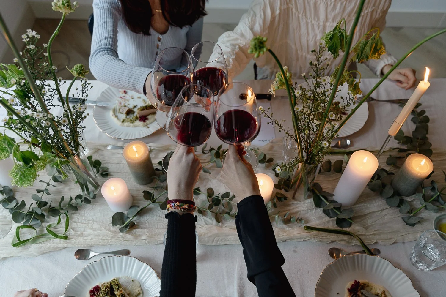 People clinking glasses of red wine at a decorated dinner table with candles and flowers.