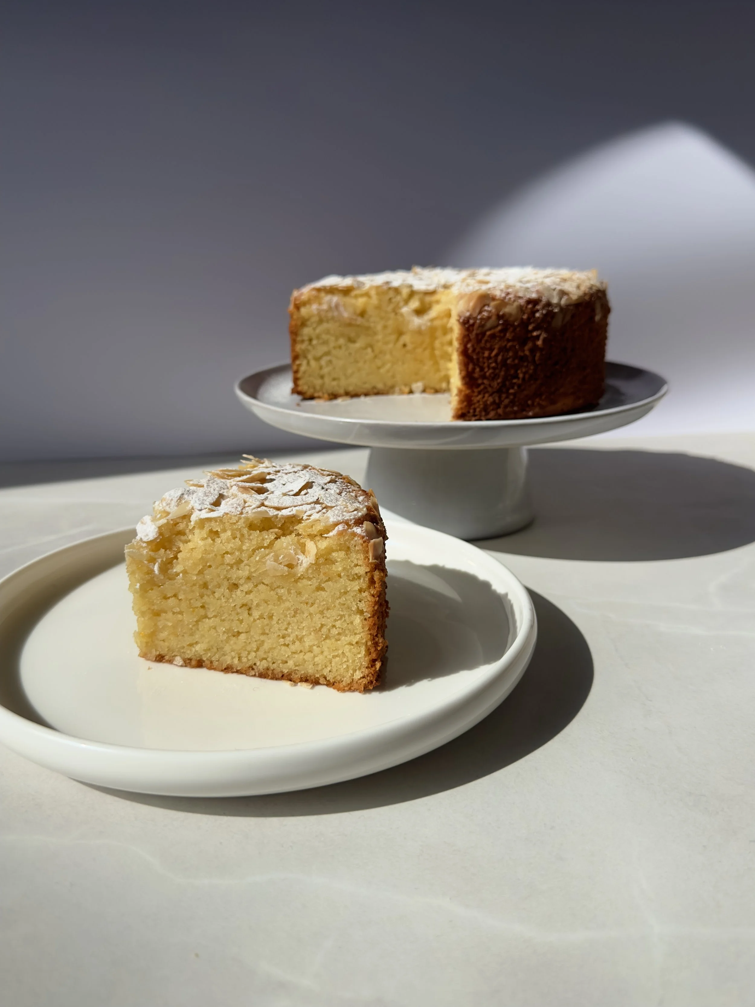A slice of sponge cake with almond slices on top on a white plate and a whole cake on a white cake stand in the background.