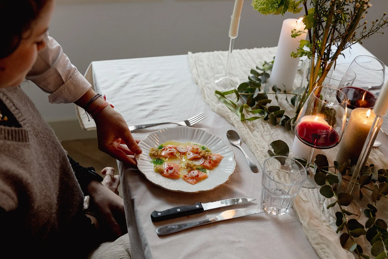 A person setting a plated dish of carpaccio with radish and microgreens on a dining table decorated with candles, greenery, and wine glasses.