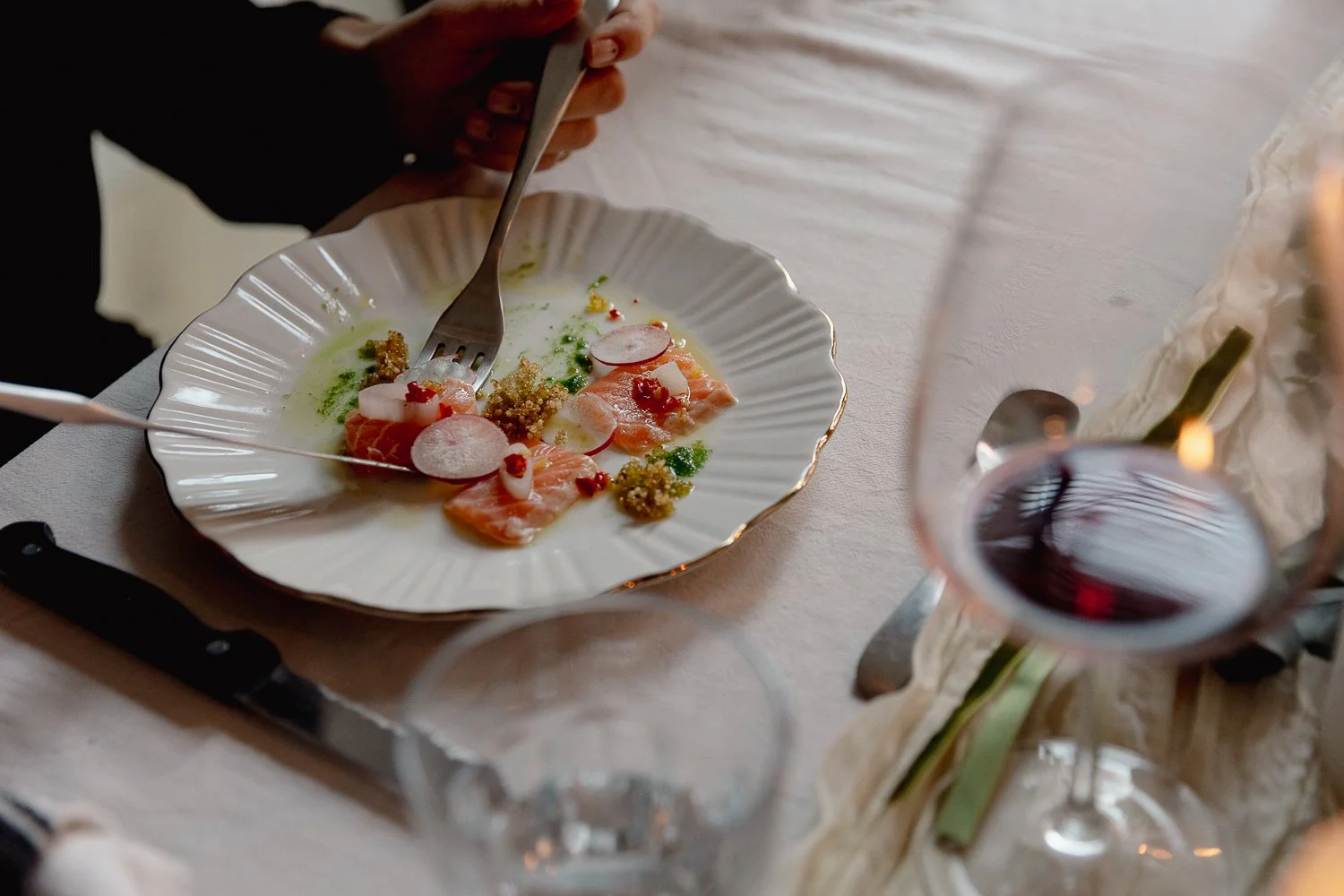 A white plate with scalloped edges holding a dish of sliced salmon, radish slices, green puree, and small red toppings, with a fork in hand, on a table with a glass of red wine, a knife, and a spoon.