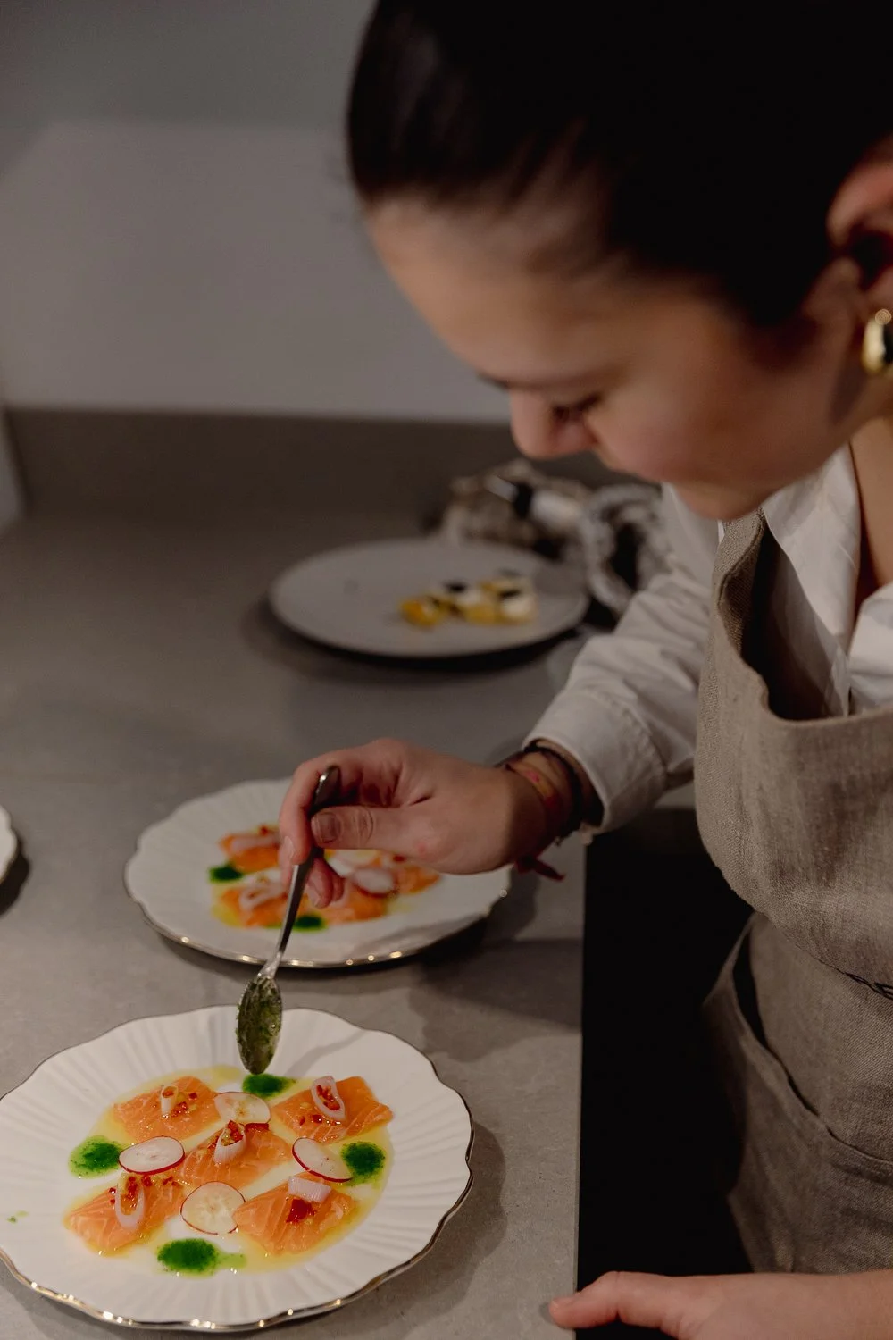 A chef is plating a dish of smoked salmon slices topped with radish slices, herbs, and sauce on white plates.