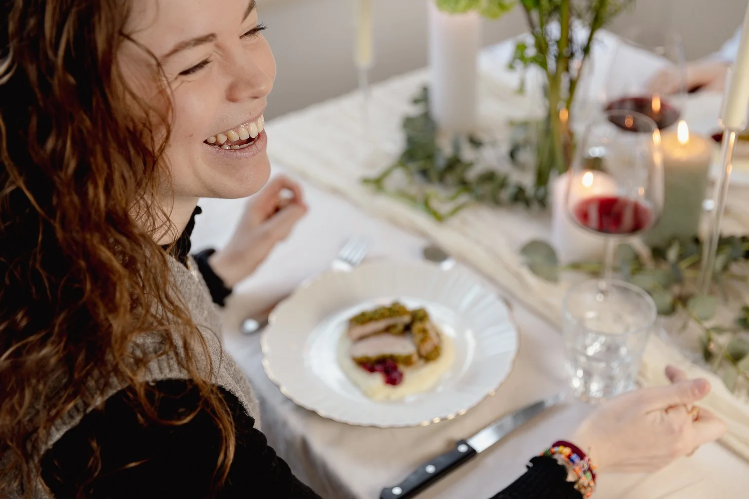 A woman with long wavy red hair enjoying a meal at a decorated dining table with glasses of red wine, candles, and greenery, with a prepared plated dish in front of her.