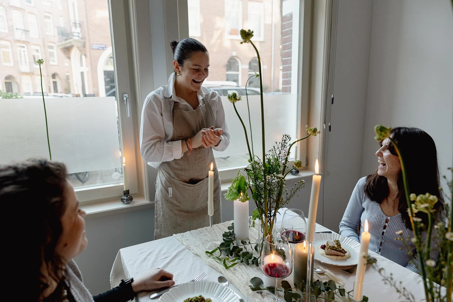 A woman in a beige apron stands and smiles at a dining table with three seated women. The table is decorated with candles, a flower arrangement, and wine glasses, with a woman holding a fork and plate of dessert.