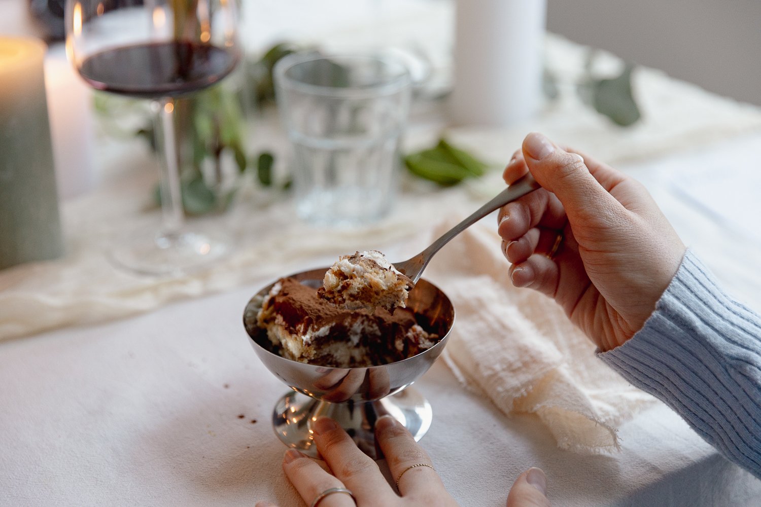 A person holding a spoonful of tiramisu in a metal dessert cup on a table, with a glass of red wine, a glass of water, and a white tablecloth in the background.