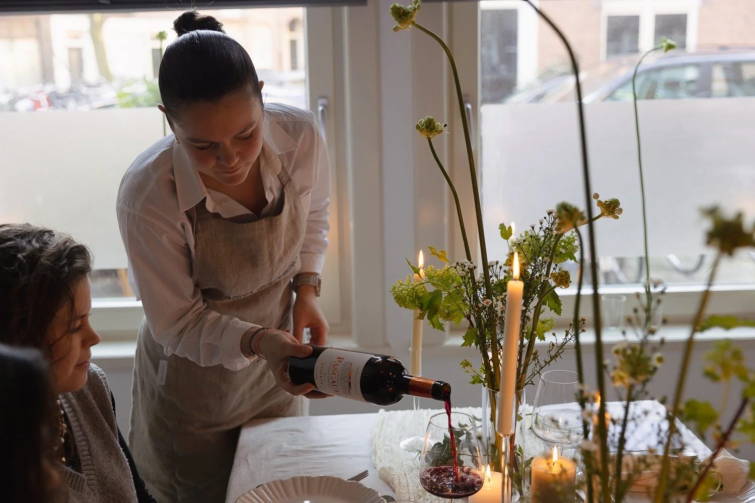 Woman pouring red wine into a glass at a table decorated with candles and flowers, with two women sitting nearby during daytime.
